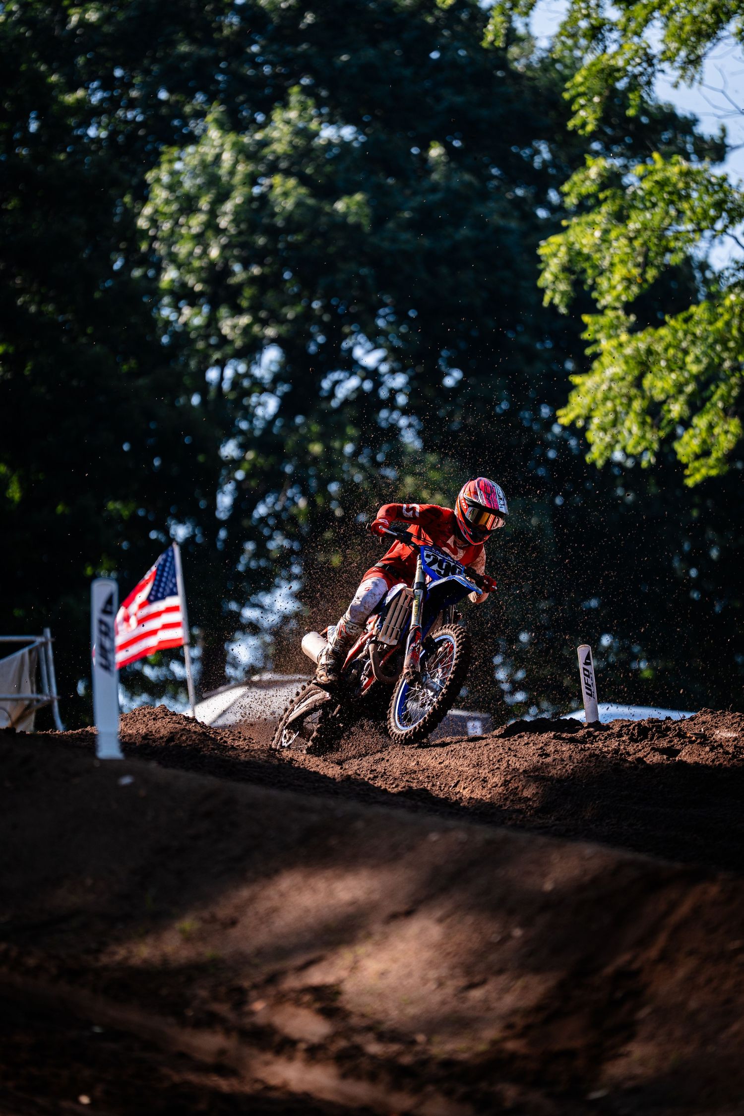 A dirt bike rider navigates a muddy track with an American flag in the background.