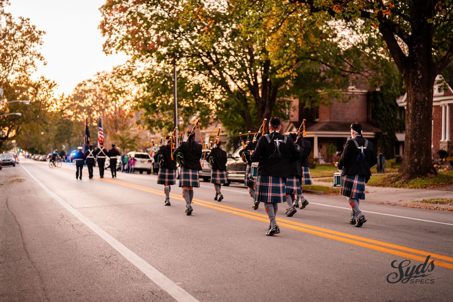 People in traditional kilts walk along a tree-lined street during an autumn parade.