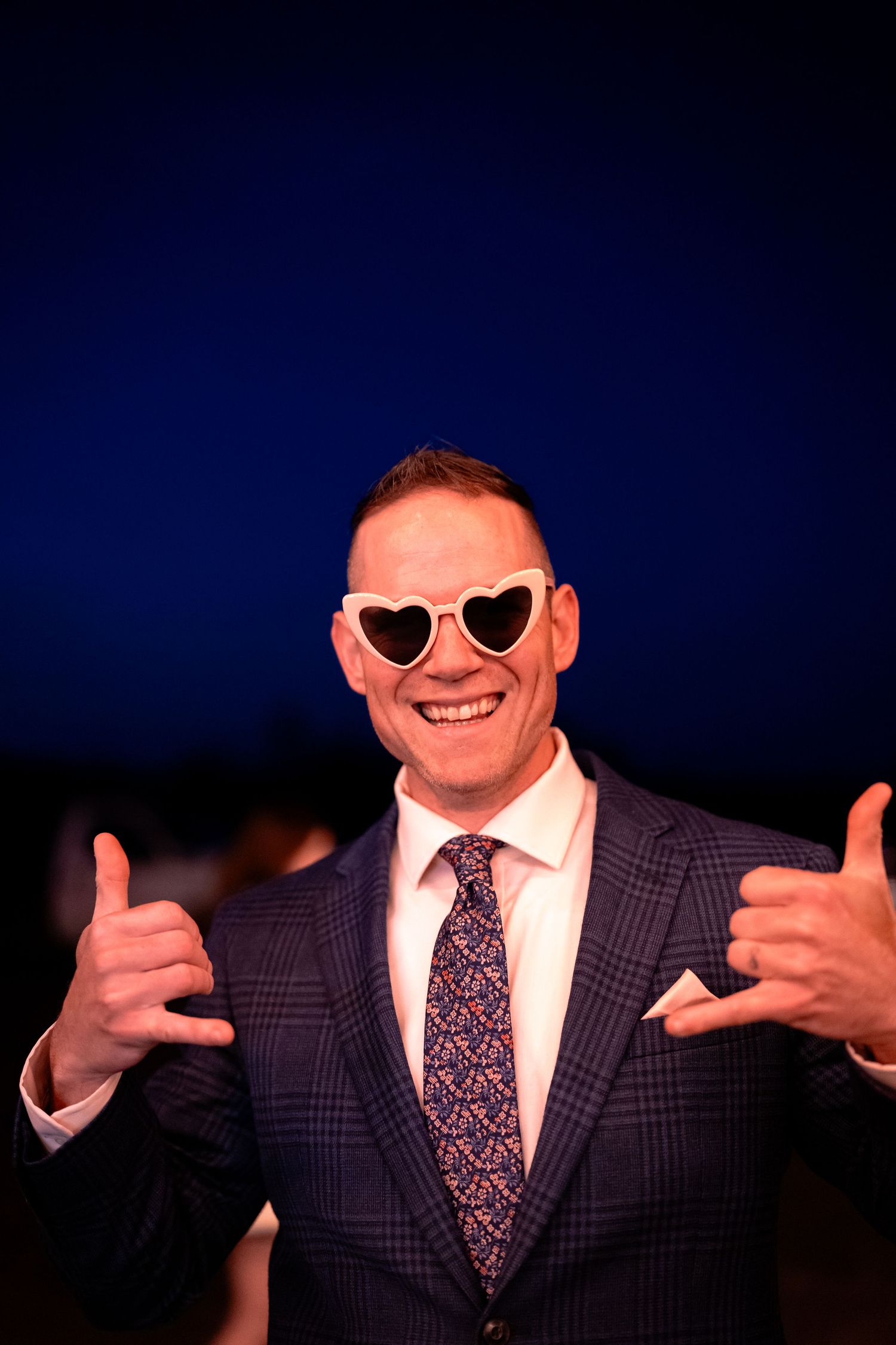 Person in formal suit wearing heart-shaped sunglasses gives thumbs up against dark blue background.