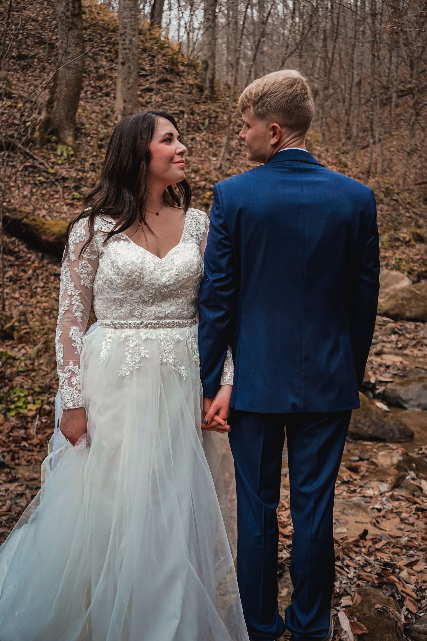 Couple in wedding attire share moment among autumn leaves in wooded setting.