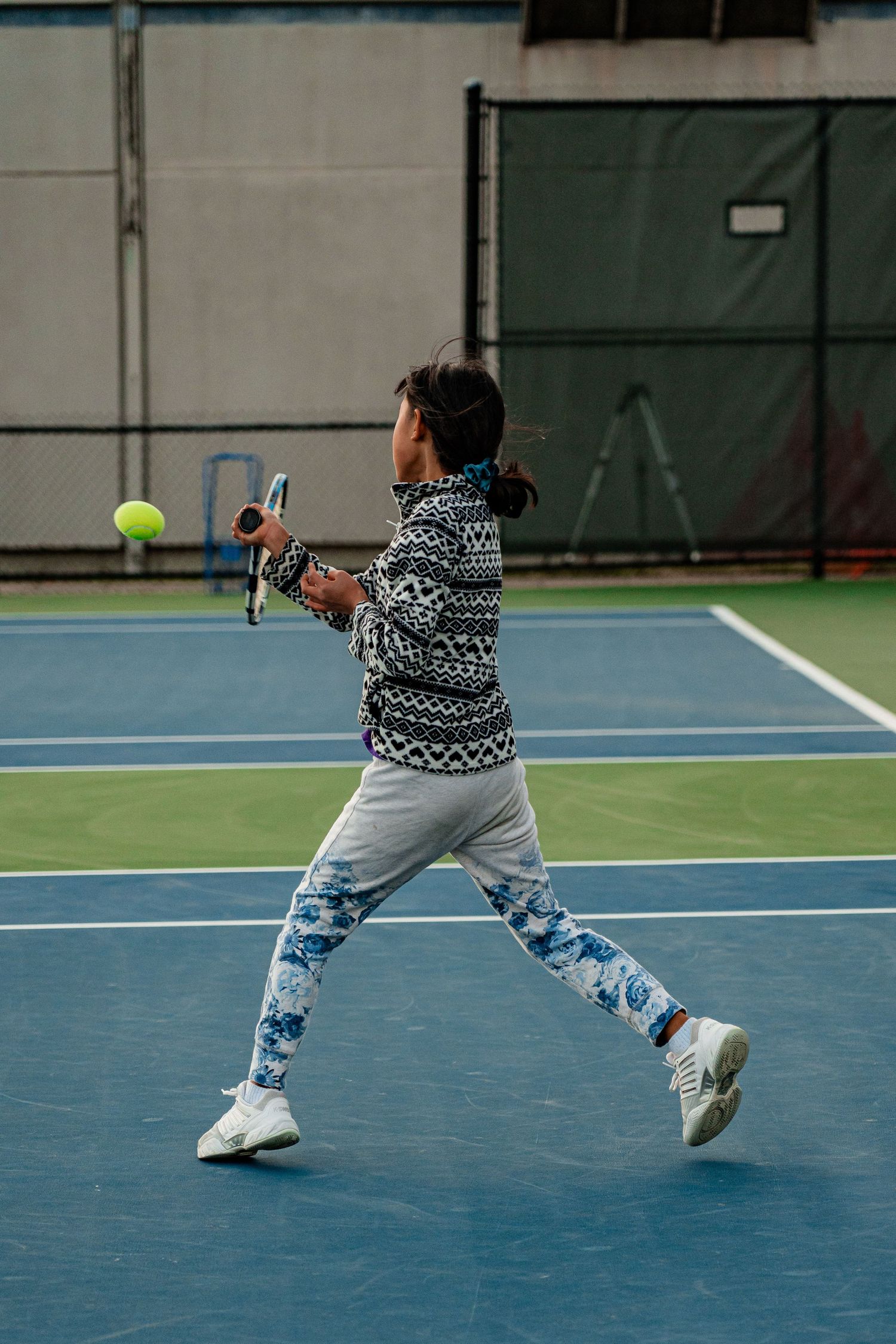 Young tennis player in patterned sweater and floral pants hits forehand shot on blue indoor court.
