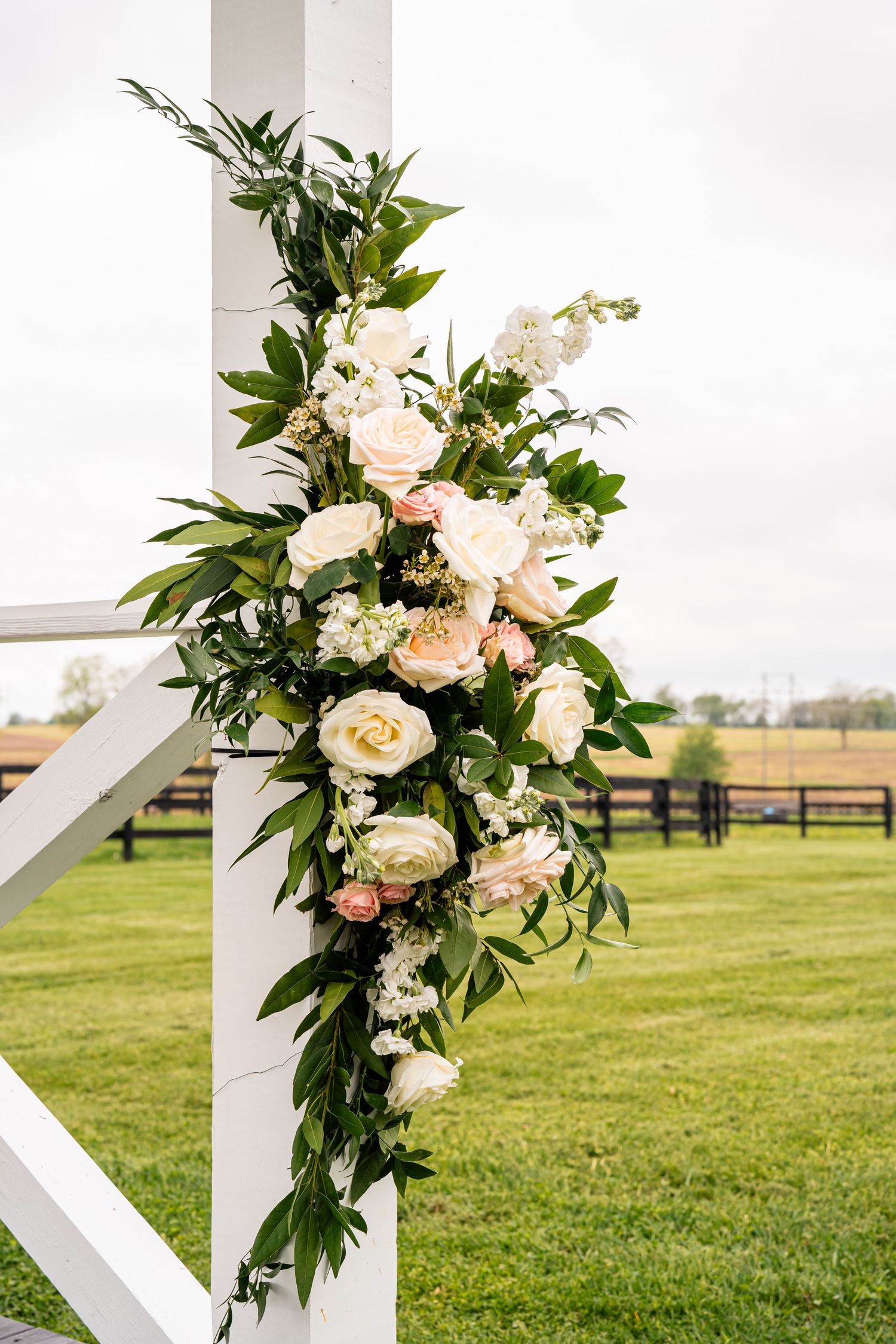Elegant white rose and greenery arrangement adorns a white post at an outdoor wedding venue.