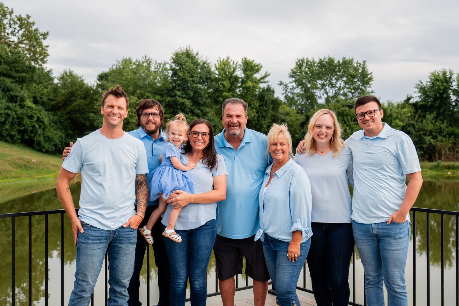 A family group wearing coordinated blue and denim outfits poses together on a deck overlooking a pond.