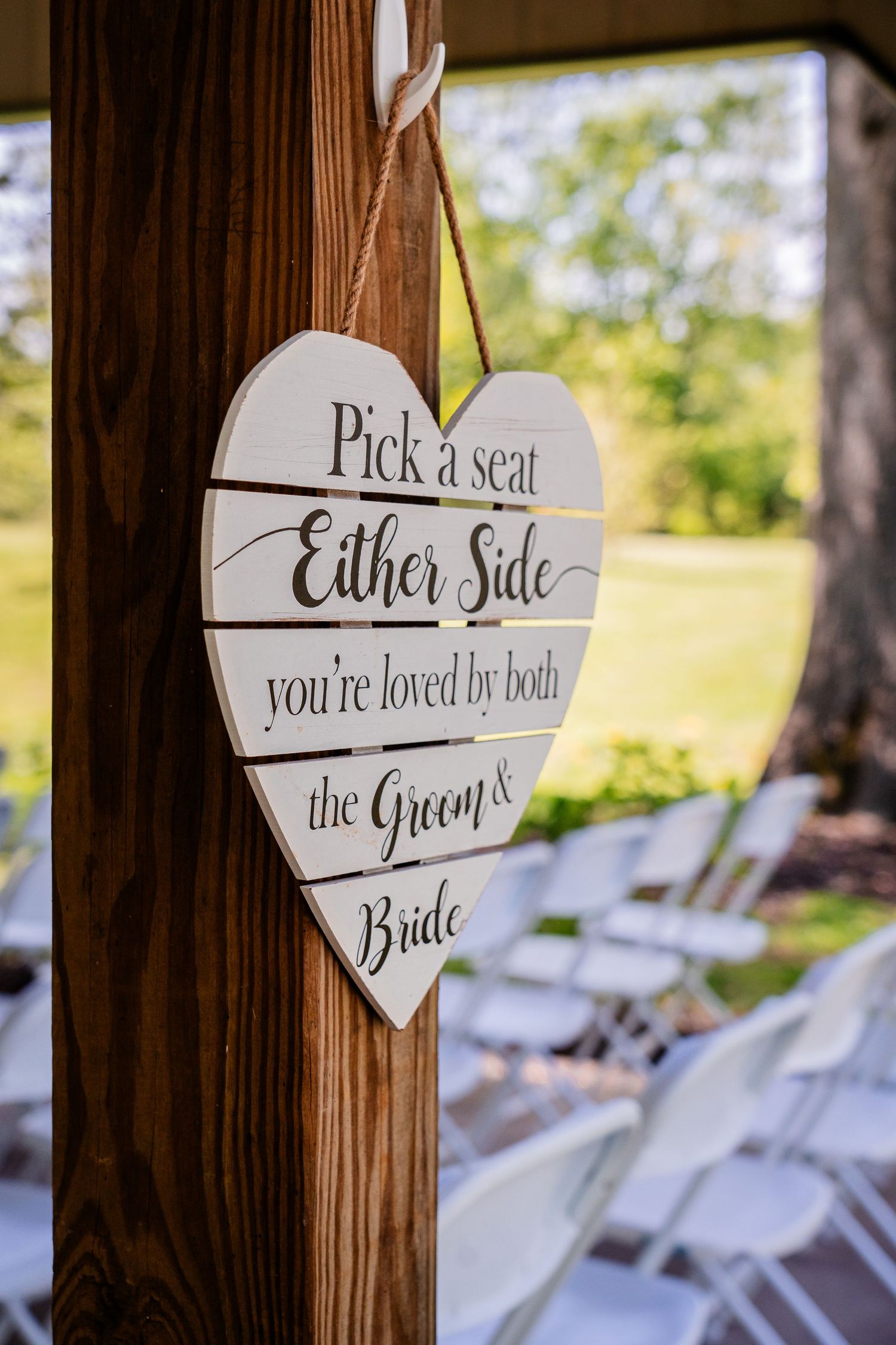 Rustic wedding seating sign reads 'Pick a seat you're loved by both' mounted on wooden post.
