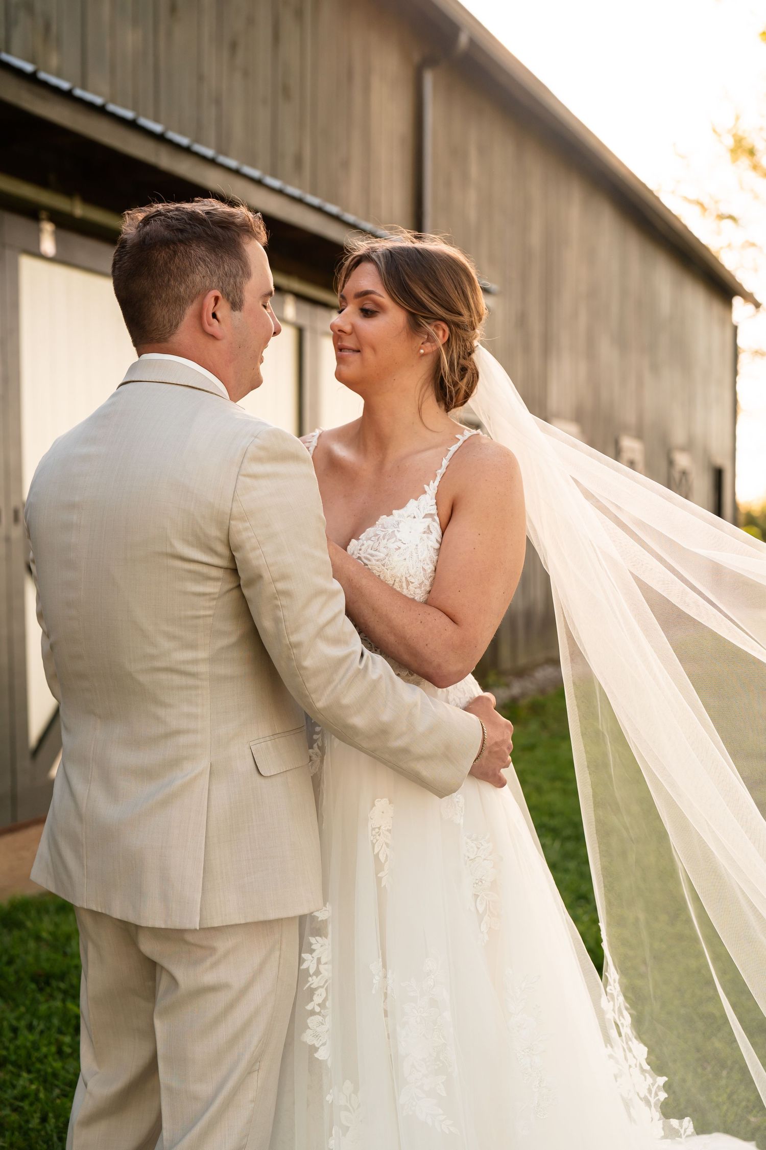 A romantic wedding couple embraces in front of a rustic barn during golden hour.