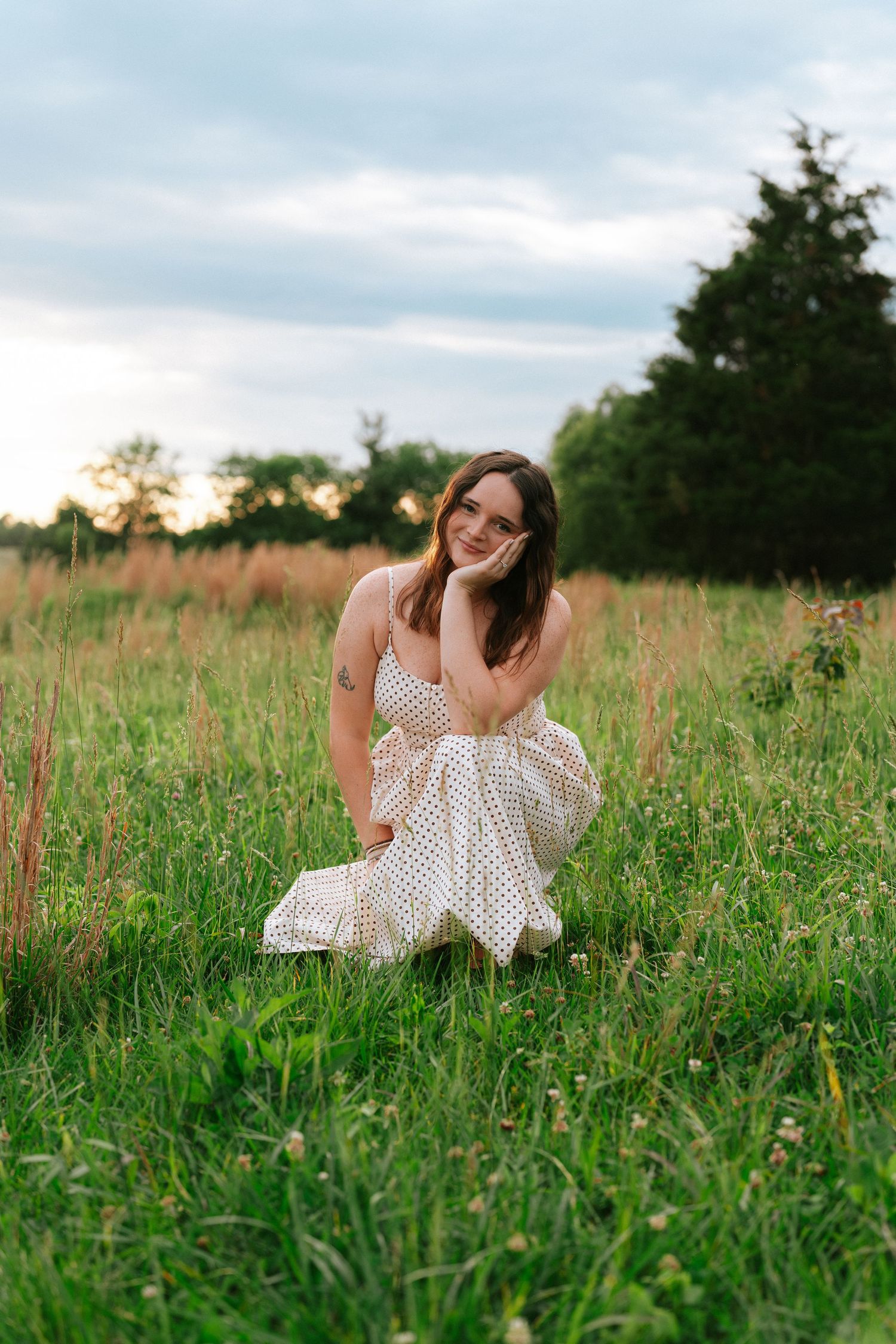 Young woman in white polka dot dress sitting in green meadow field smiling at sunset.
