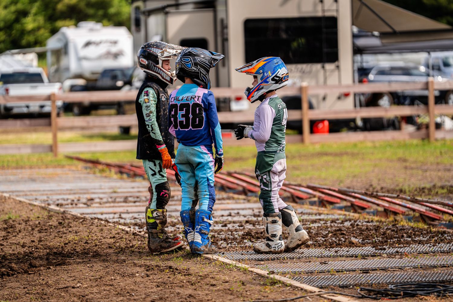 Motocross riders prepare at starting line of dirt track with support crew nearby.