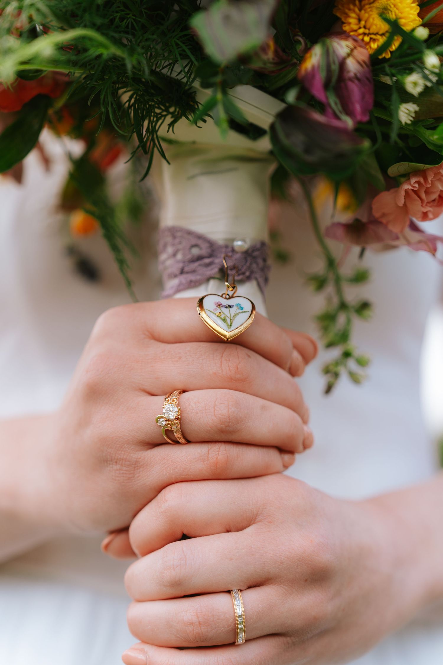Bride holding colorful wedding bouquet with heart locket charm, wearing gold diamond engagement ring and wedding band.