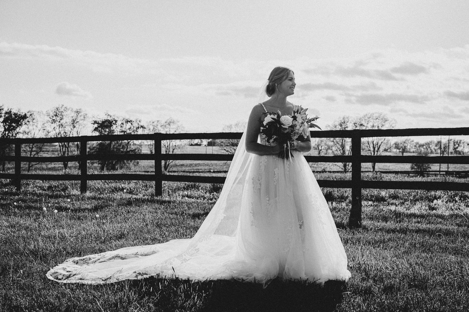 A flowing white wedding dress train trails across a rustic wooden fence in an outdoor setting.