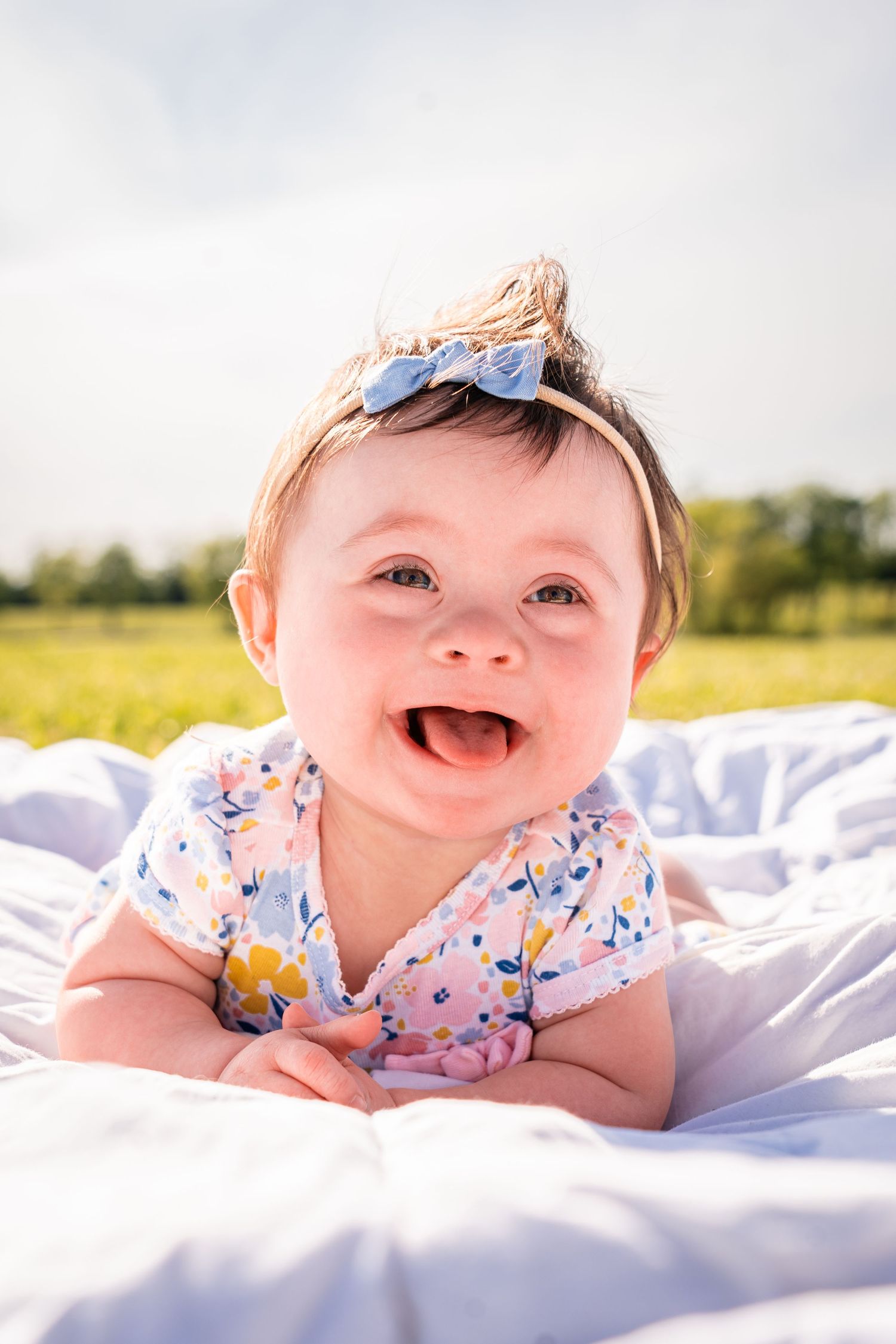 A happy baby wearing floral print and blue headband lies on white blanket outdoors.