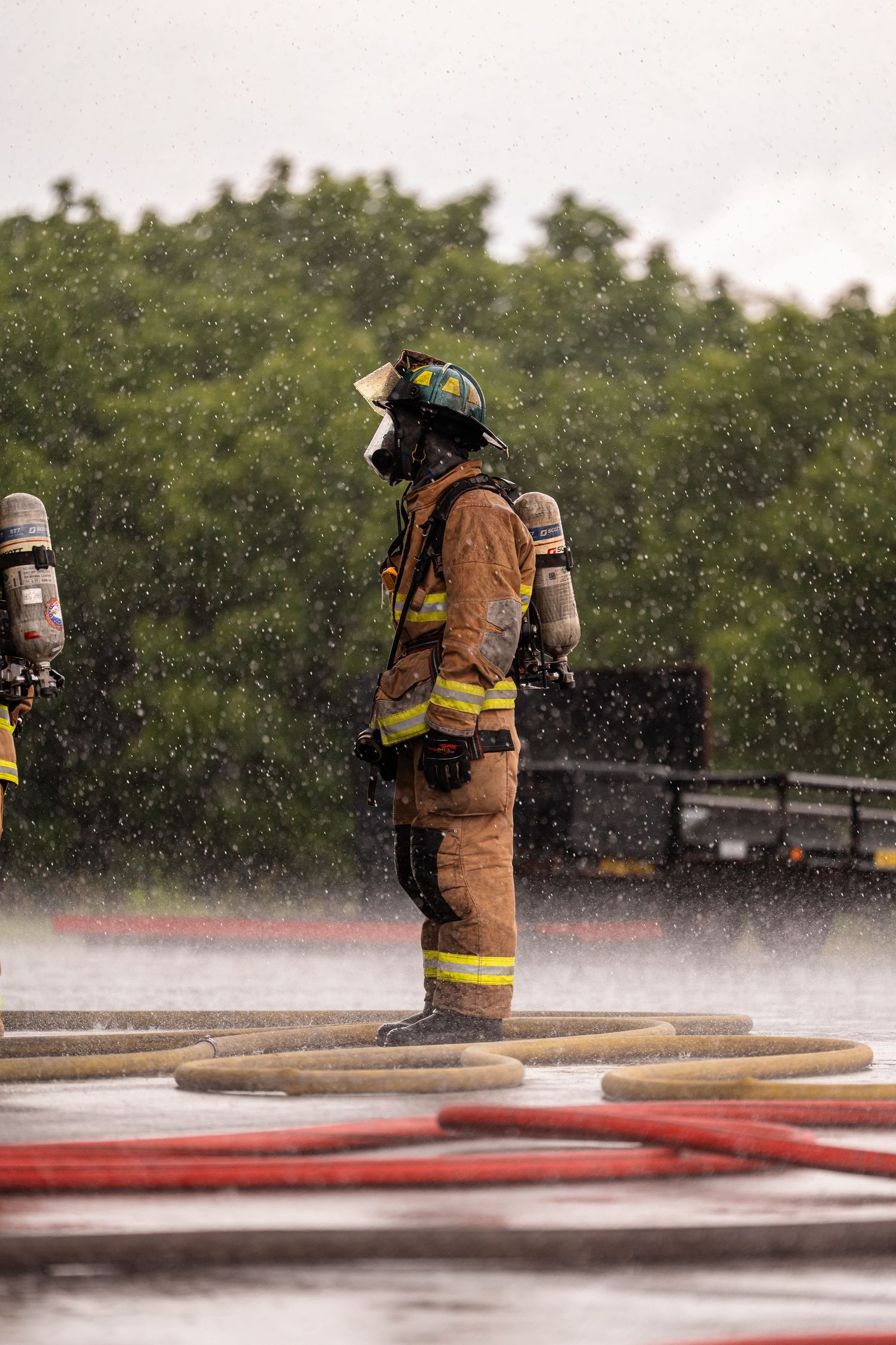 A firefighter stands in the rain during a training exercise with fire hoses visible.