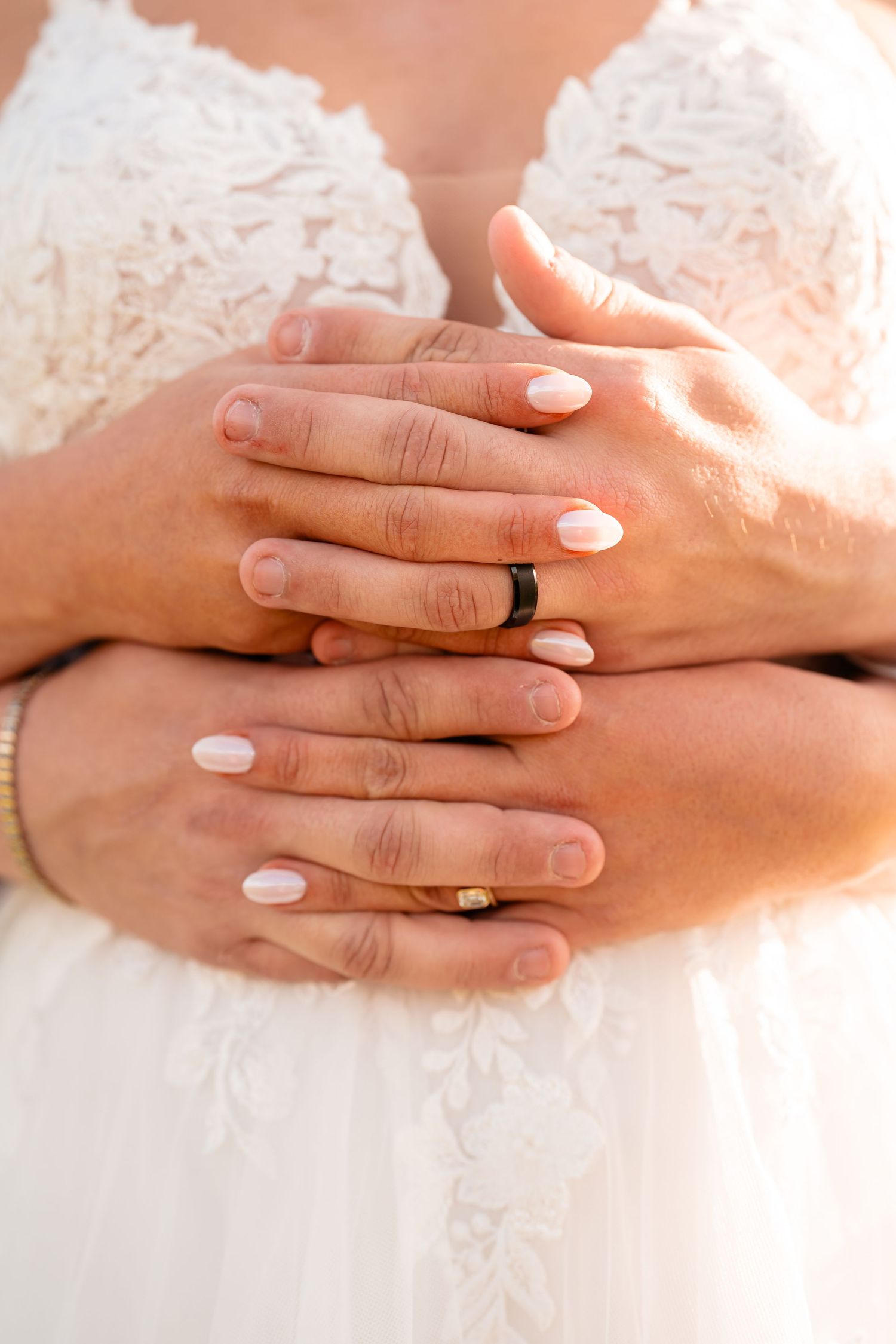 Close-up of hands with wedding rings and white manicure against lace dress.