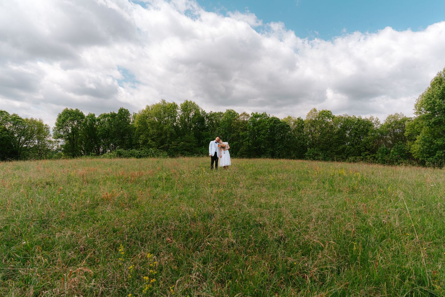 Couple embracing in a lush green meadow with wildflowers, trees, and dramatic cloudy sky in background.