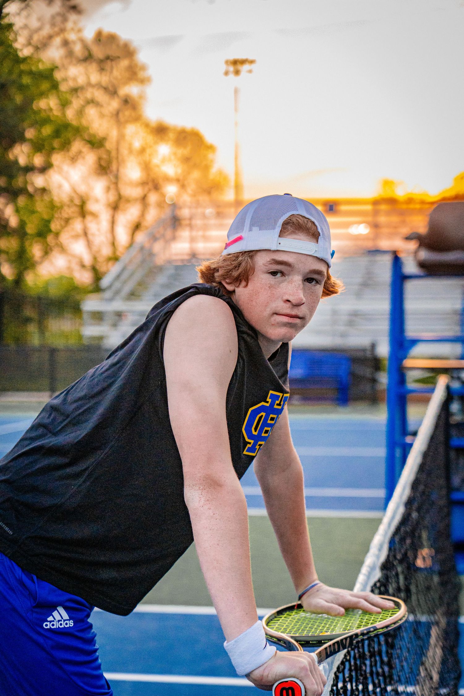 Tennis player in sleeveless shirt leans on net during sunset practice session.