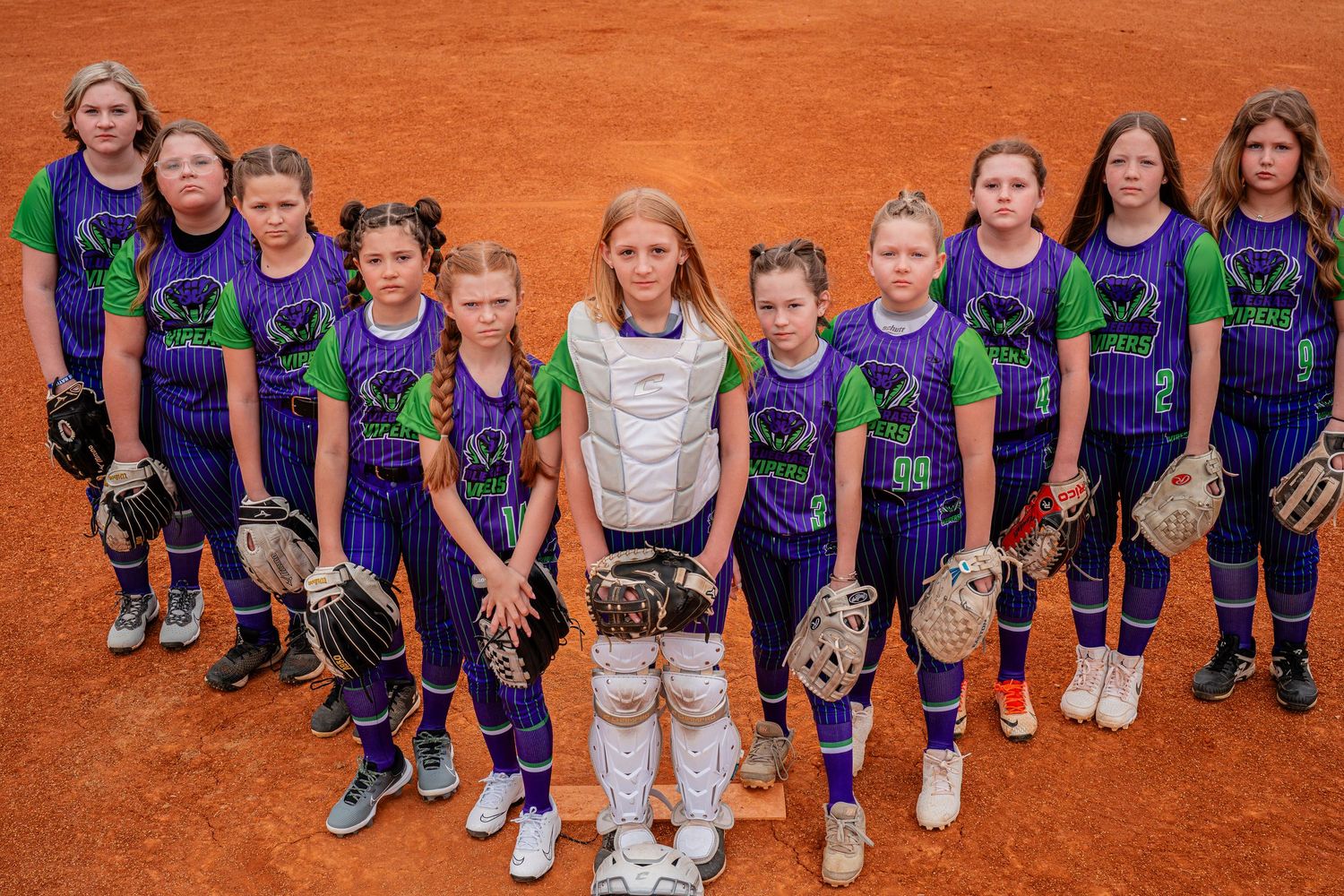 Youth softball team in purple and green striped uniforms poses together on dirt field.