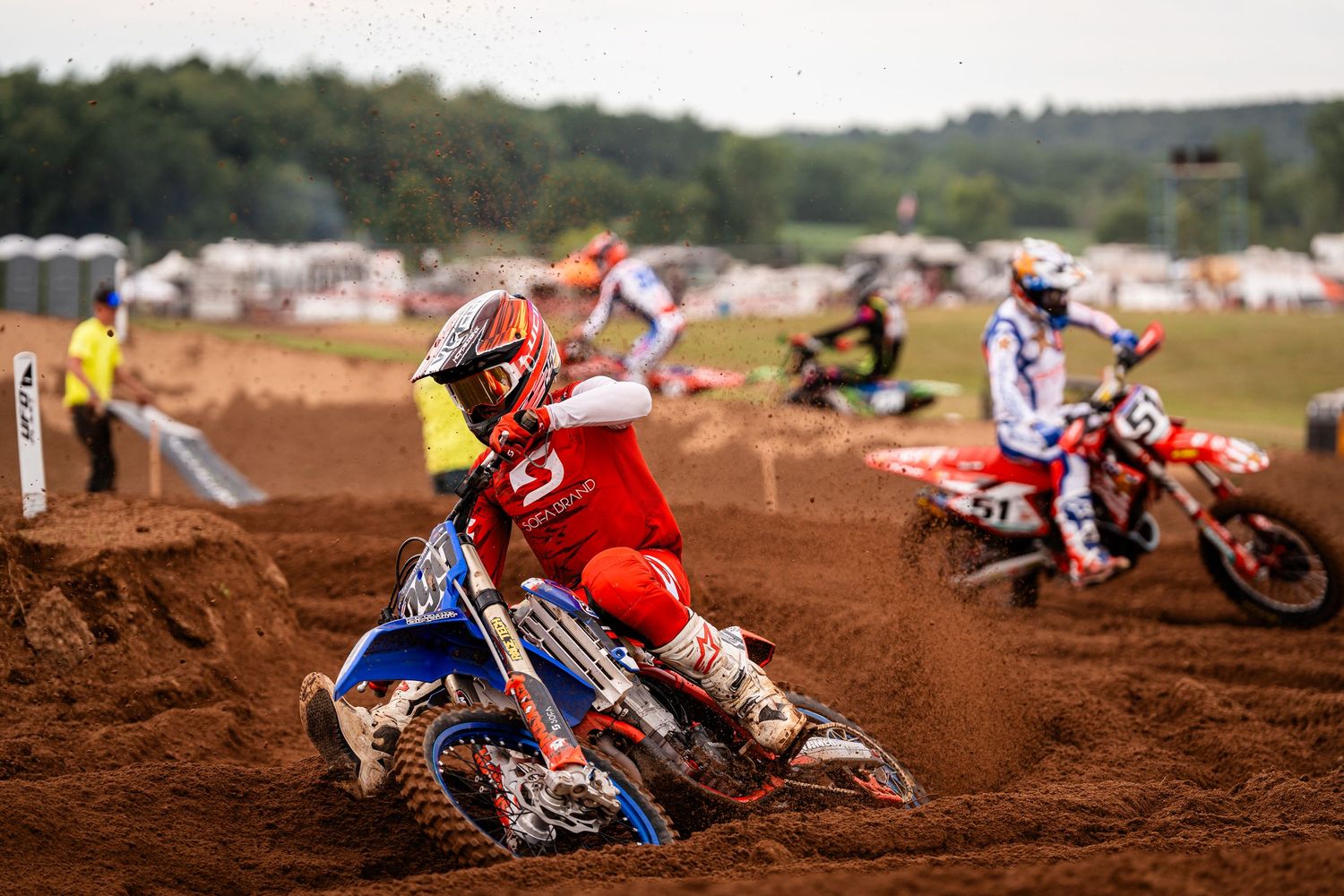 Motocross riders racing on a dirt track surrounded by spectators and tents in the background.