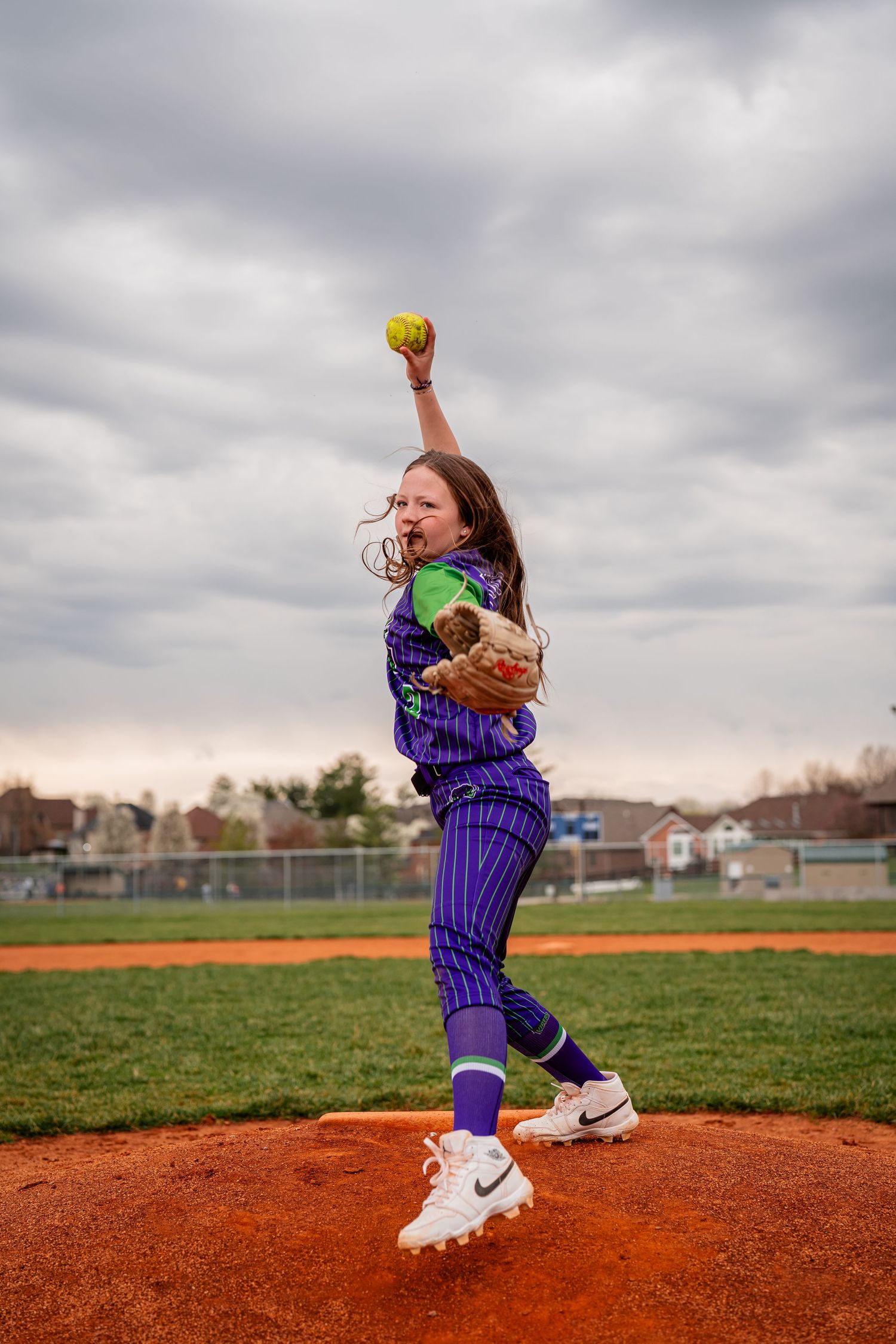 Young softball player in purple uniform practicing pitching technique on dirt mound.