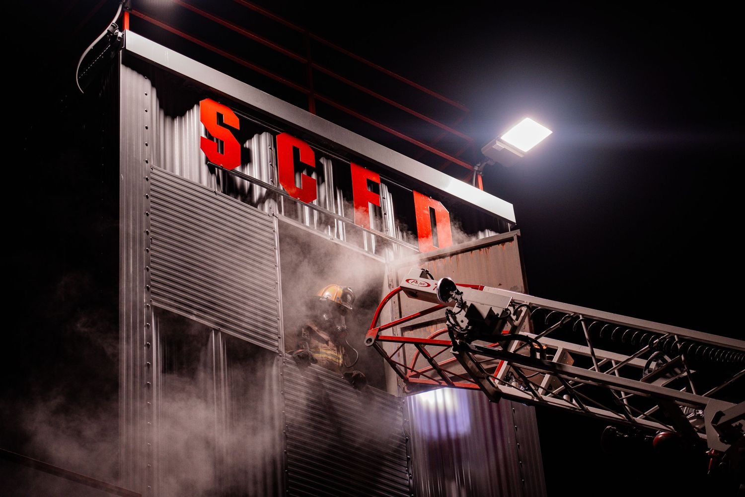 Firefighters on a ladder truck battle a blaze at an SCFD building at night, with smoke and dramatic lighting.