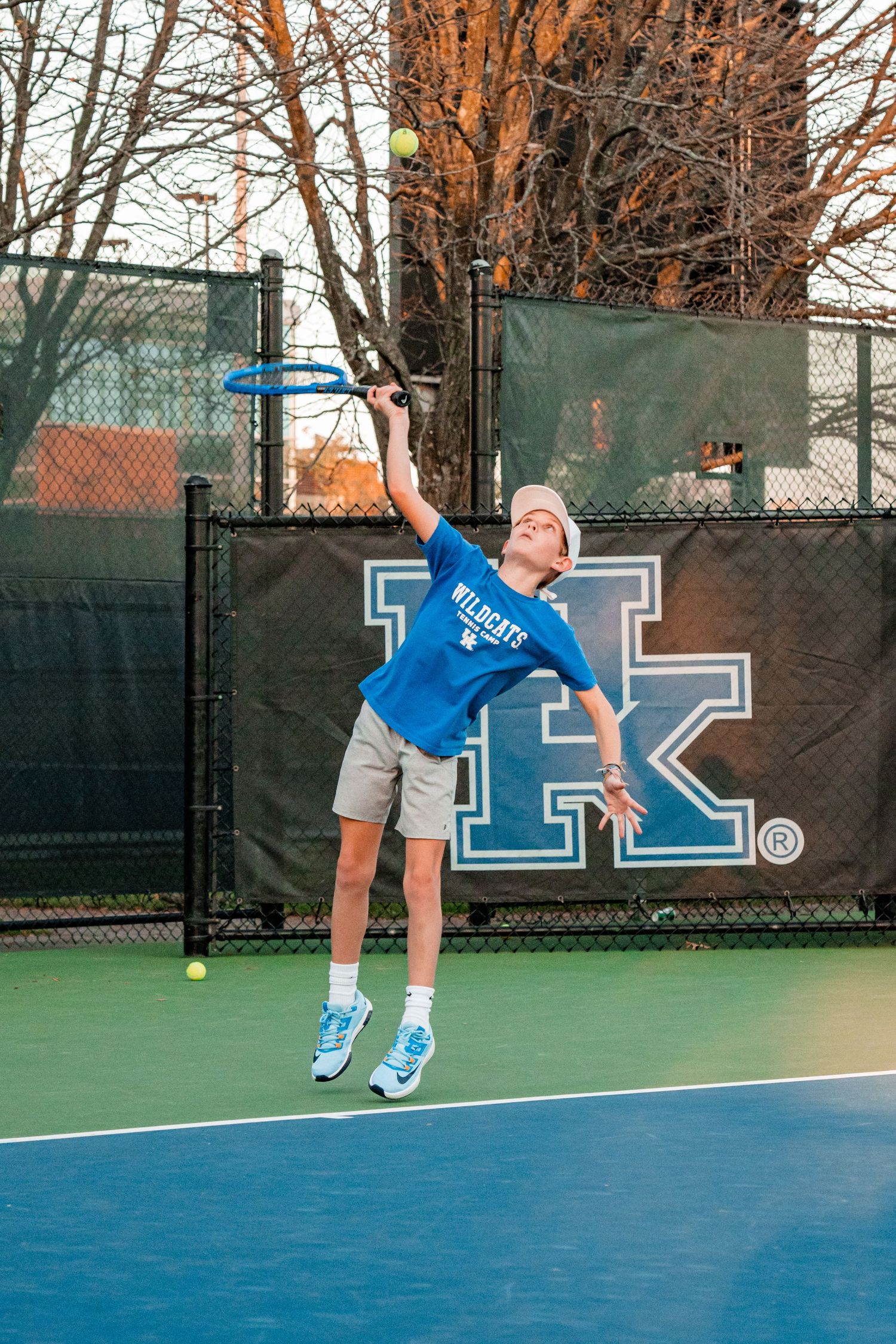 Tennis player in blue shirt serves ball on outdoor court with Kentucky logo visible.