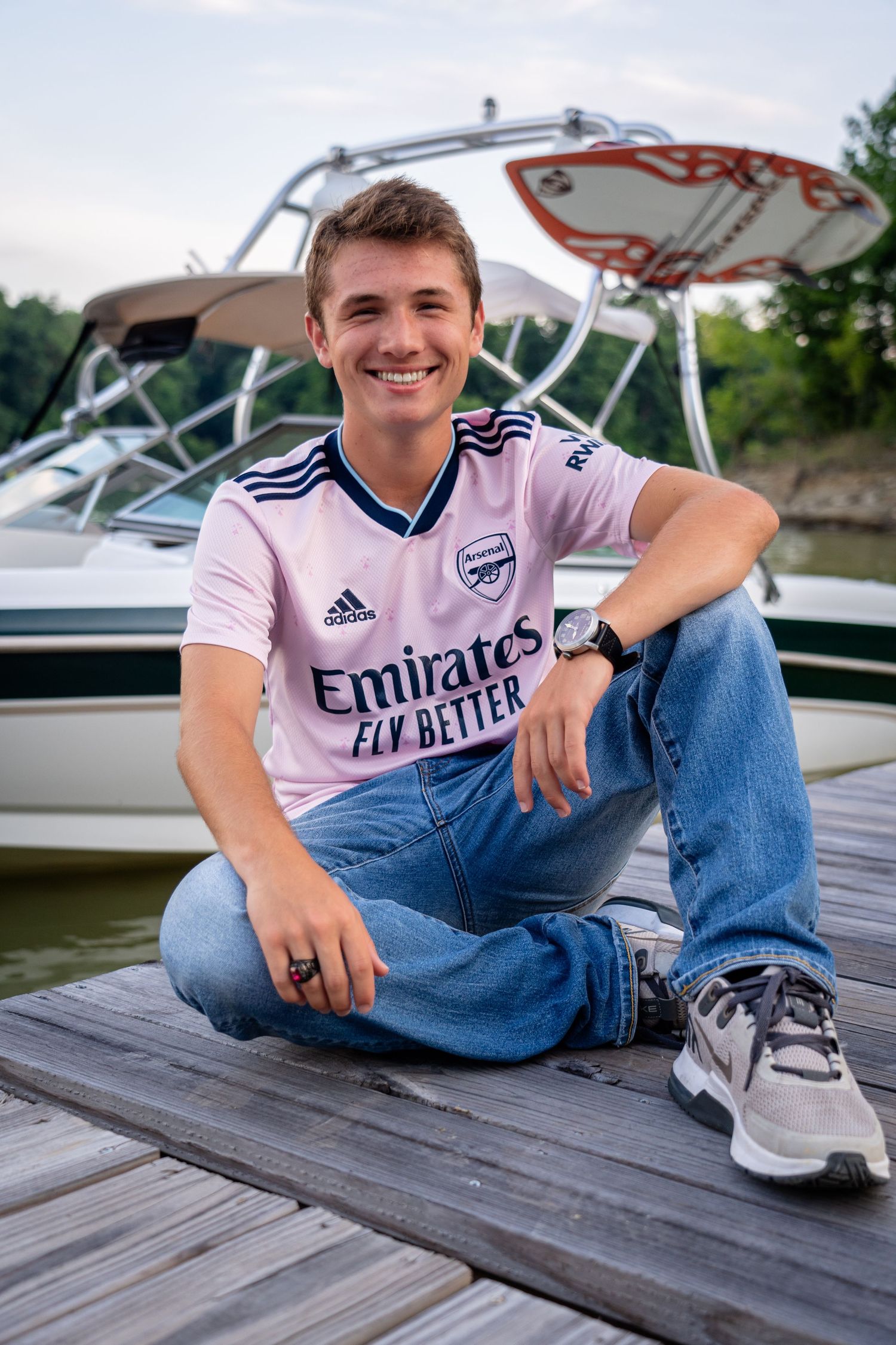 Person wearing an Arsenal soccer jersey sits casually on wooden dock by water.