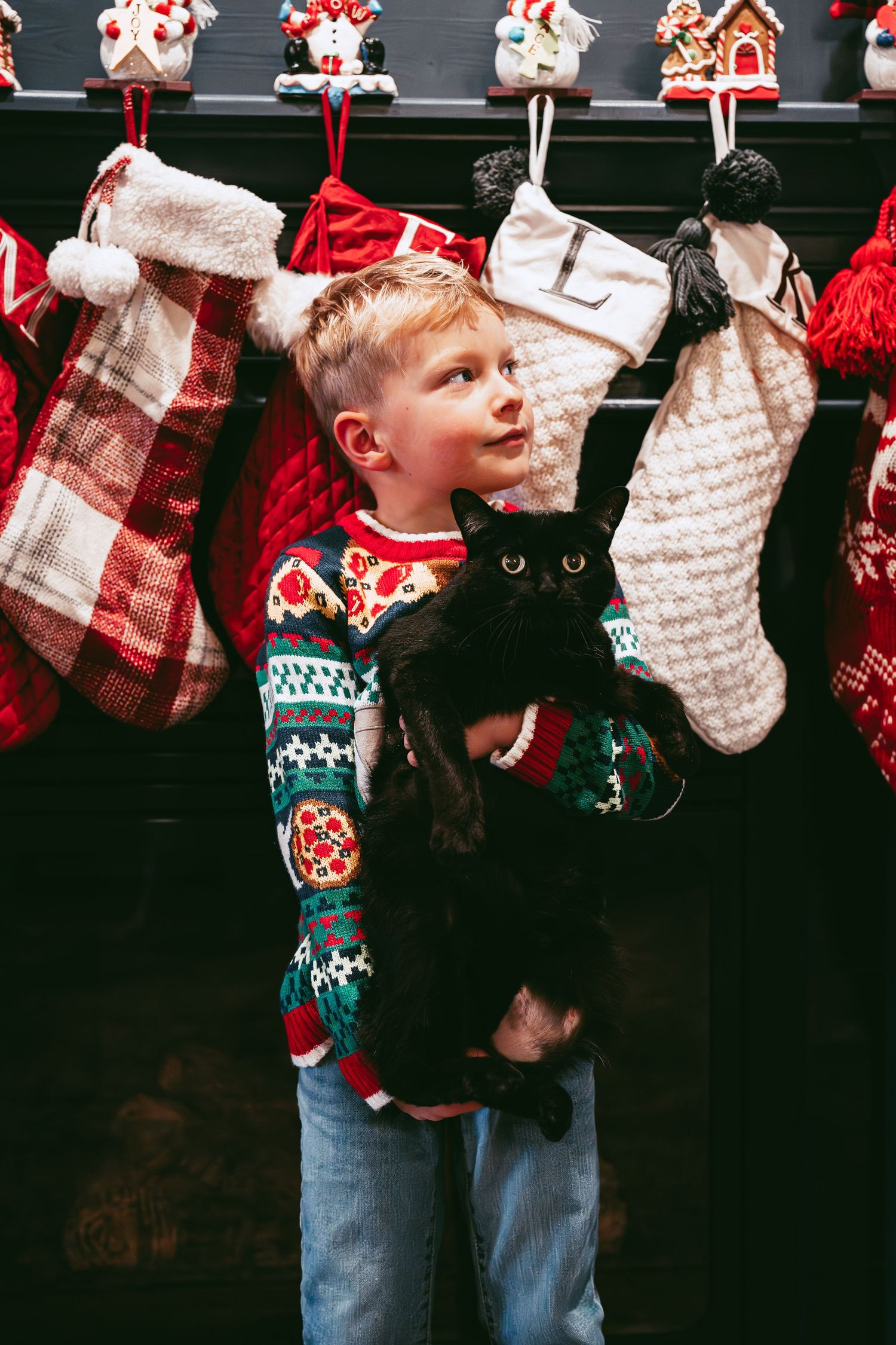 A young child in a festive sweater stands by hanging Christmas stockings and holiday decorations.