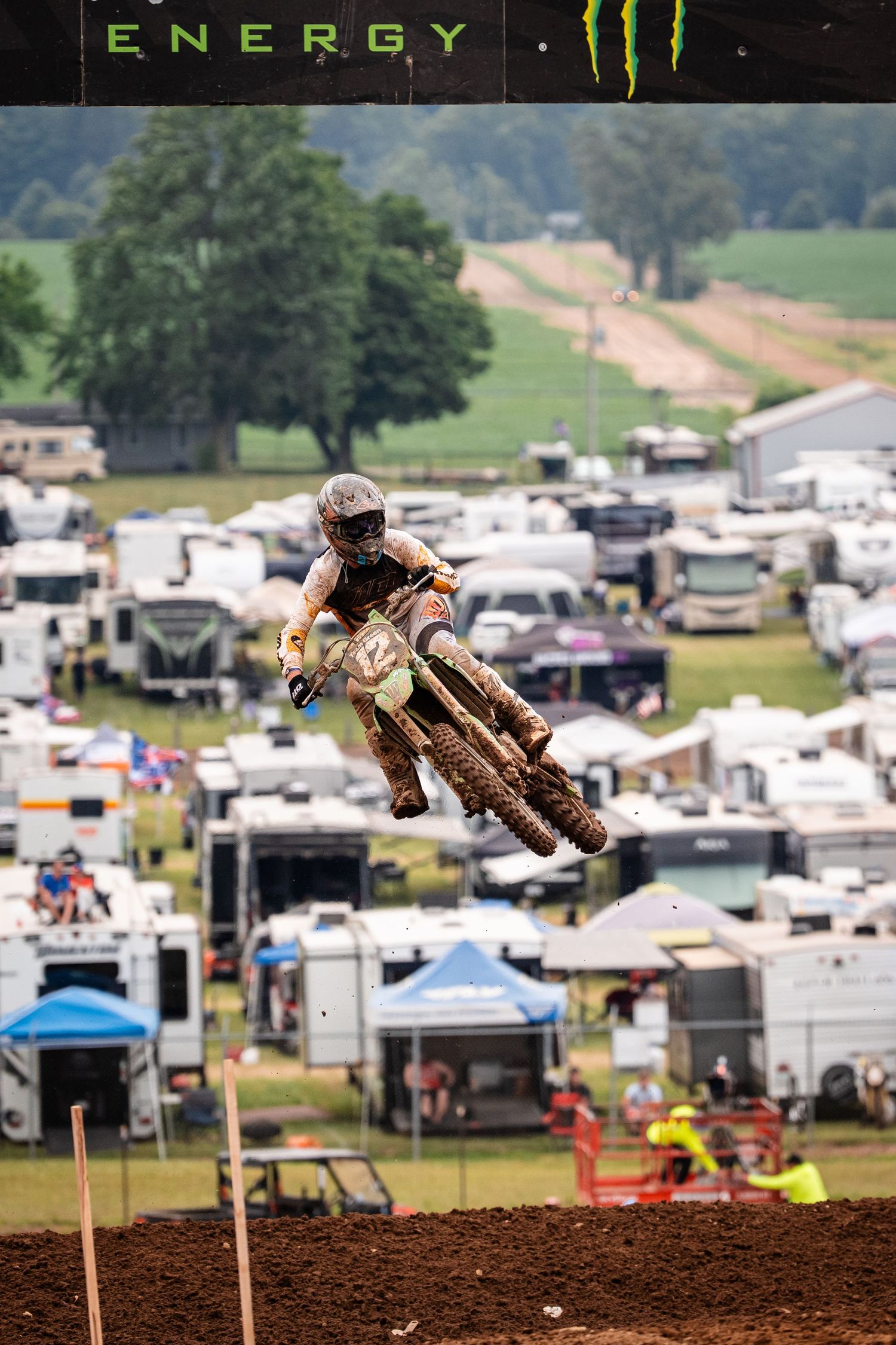 Motocross rider soaring through air over dirt jump with RV camping area in background.