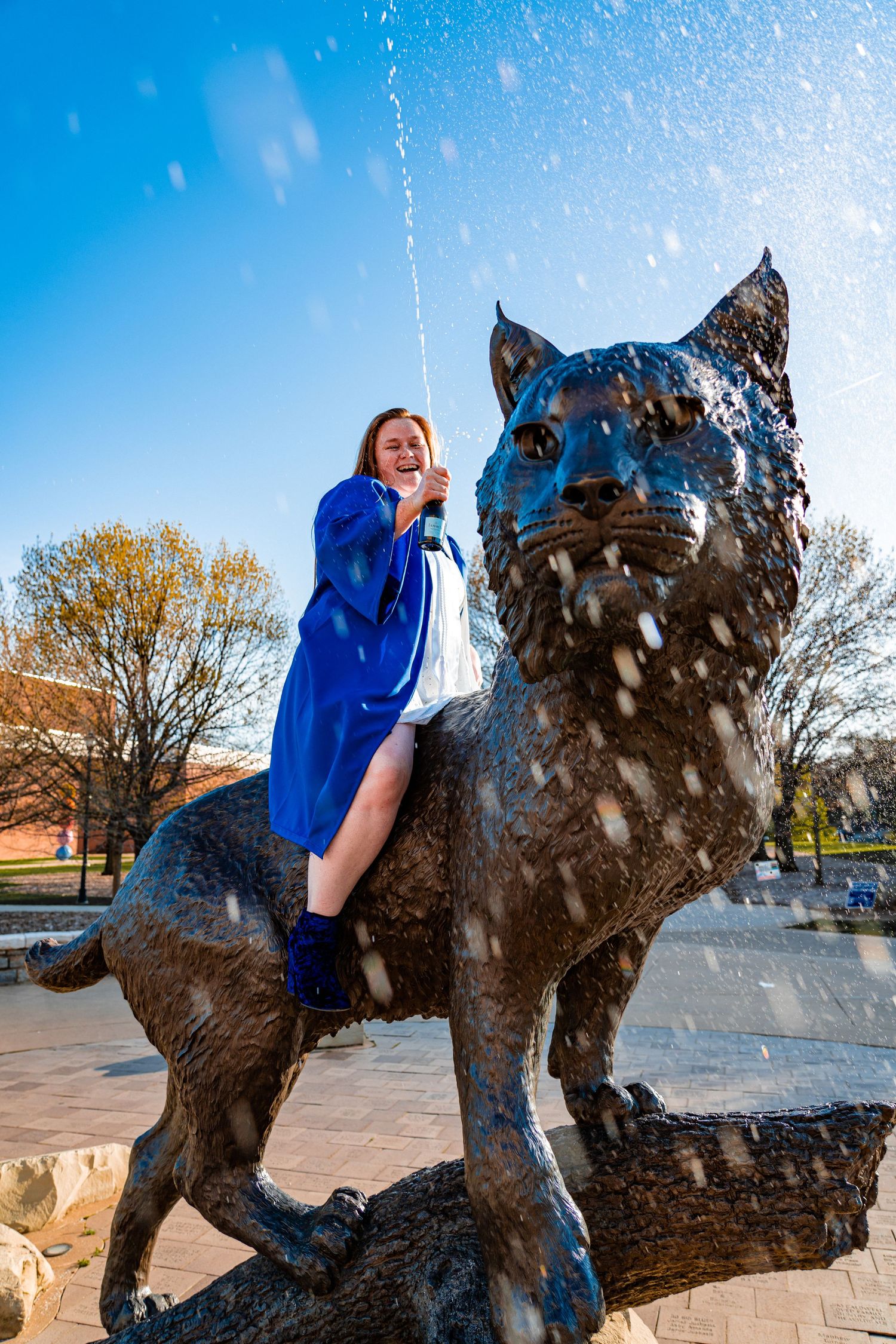 Graduate in blue gown poses playfully on wildcat statue with water splashing.