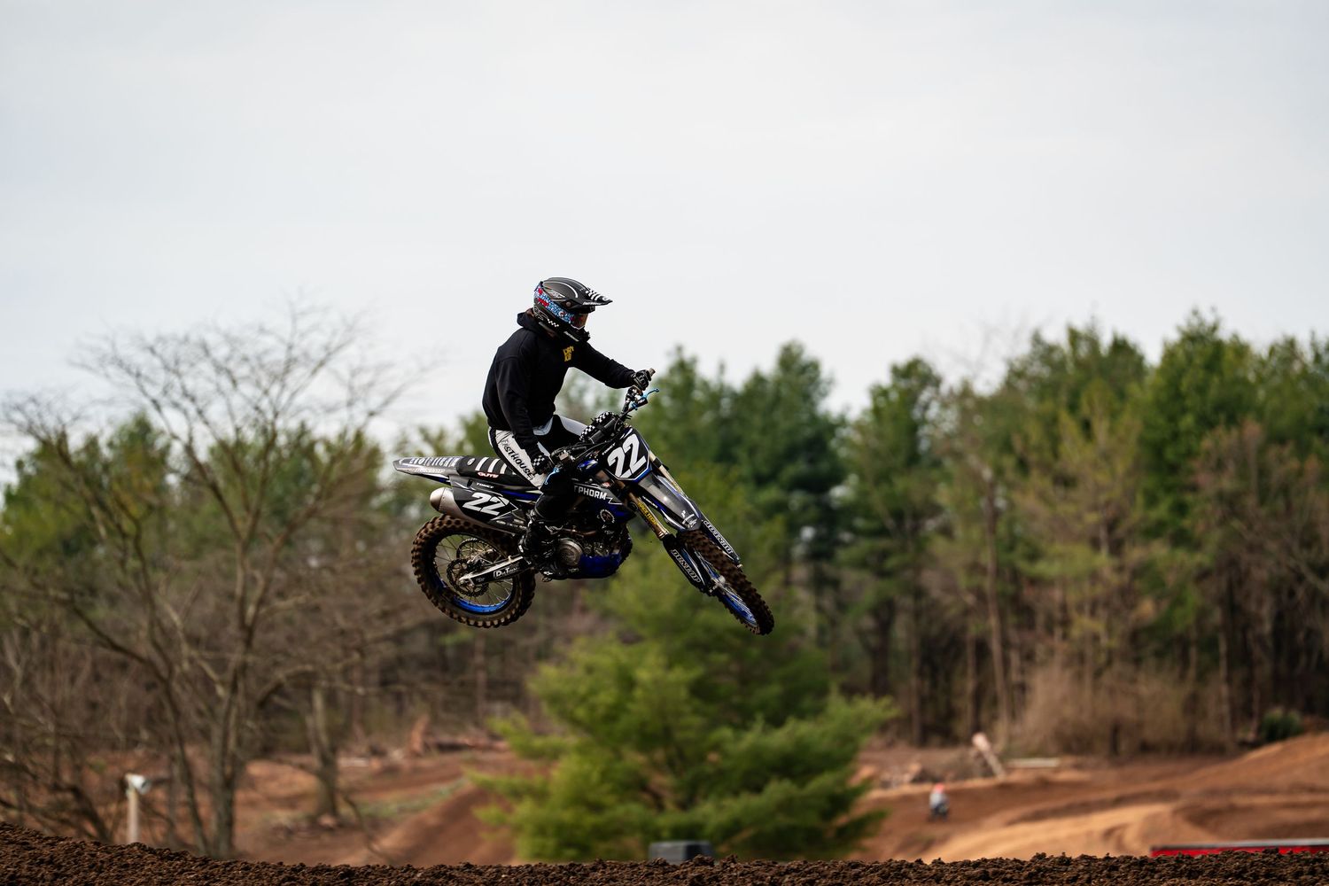 A person riding a dirt bike jumps over a dirt track surrounded by trees.