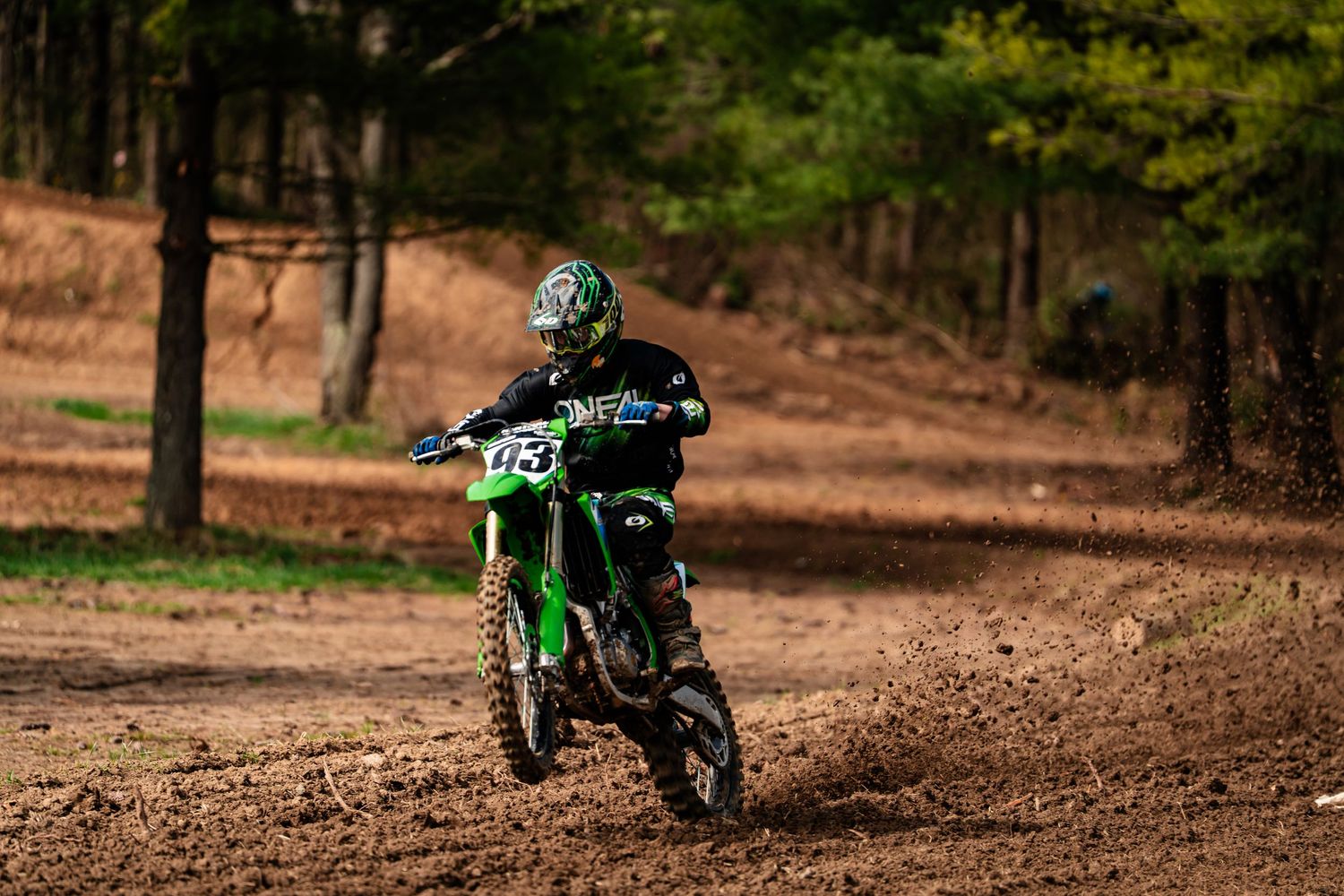 A person riding a green off-road motorcycle on a dirt trail surrounded by trees.