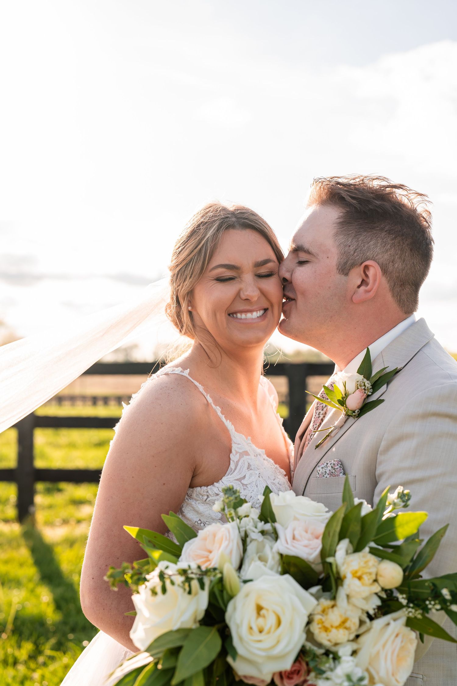 A tender moment between newlyweds with white roses and greenery bouquet against a countryside backdrop.