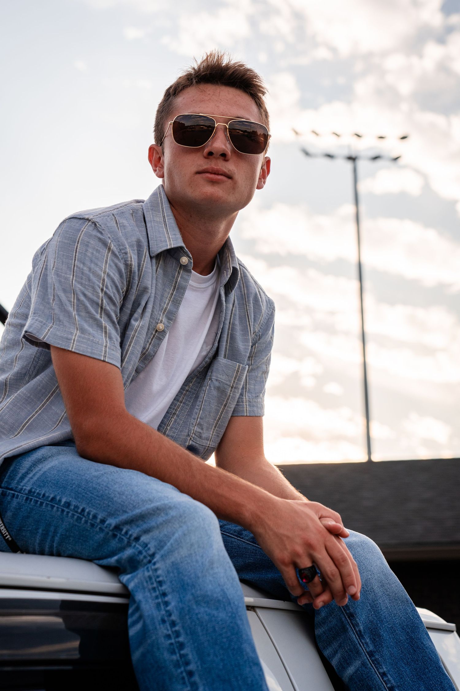 Person in plaid shirt and jeans relaxing on car hood at sunset.
