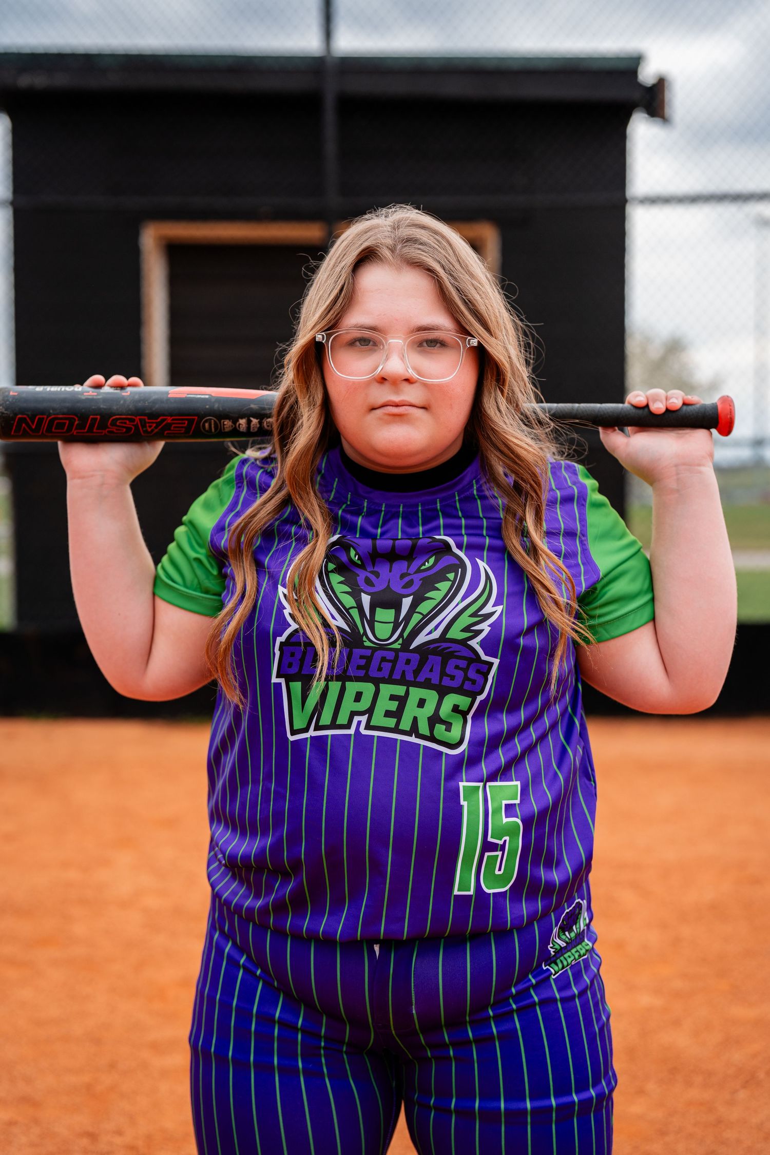 Softball player in purple Vipers jersey holds bat across shoulders on dirt field.