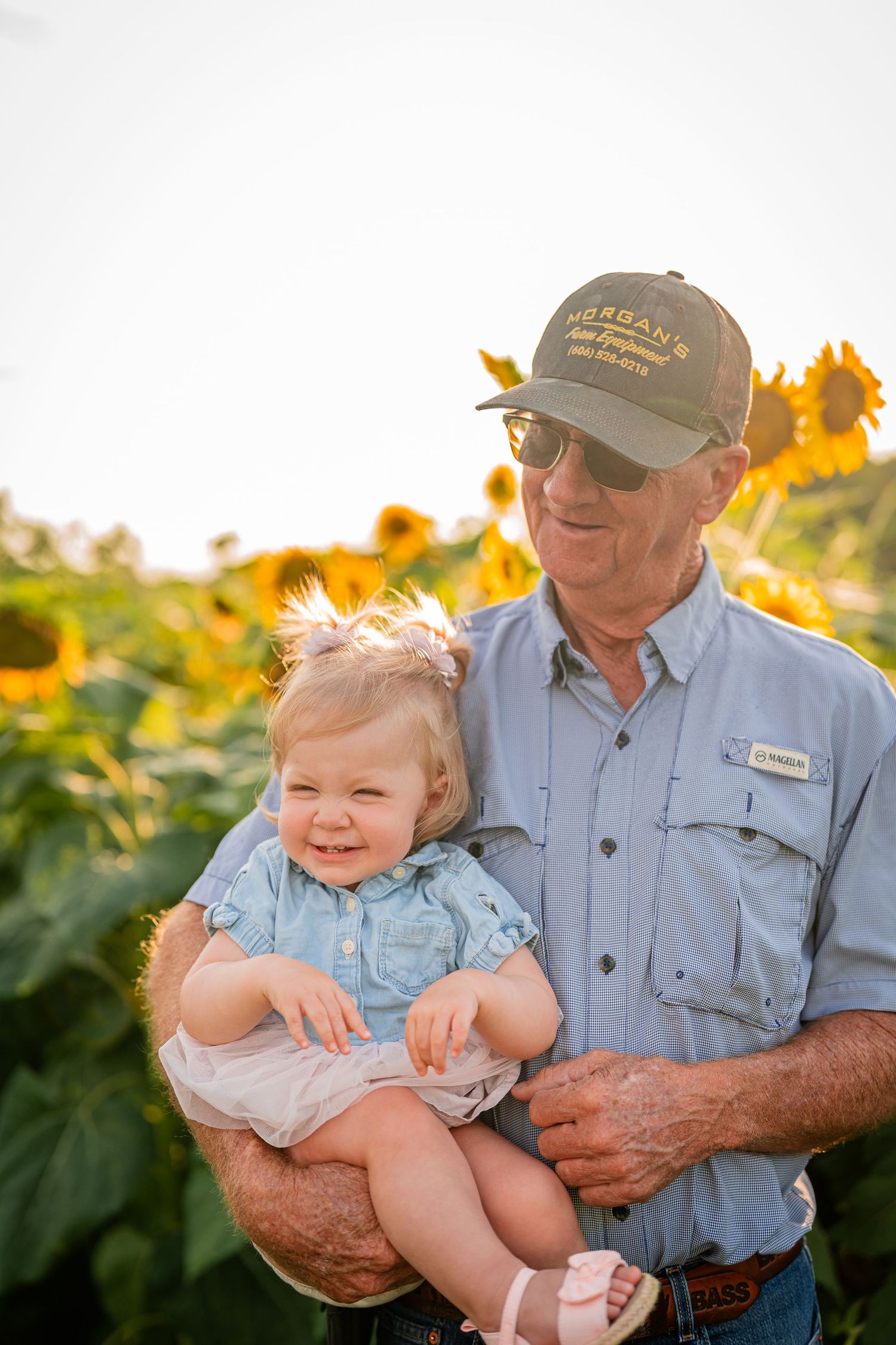 Elderly farmer in work clothes sharing special moment with baby in sunflower field.