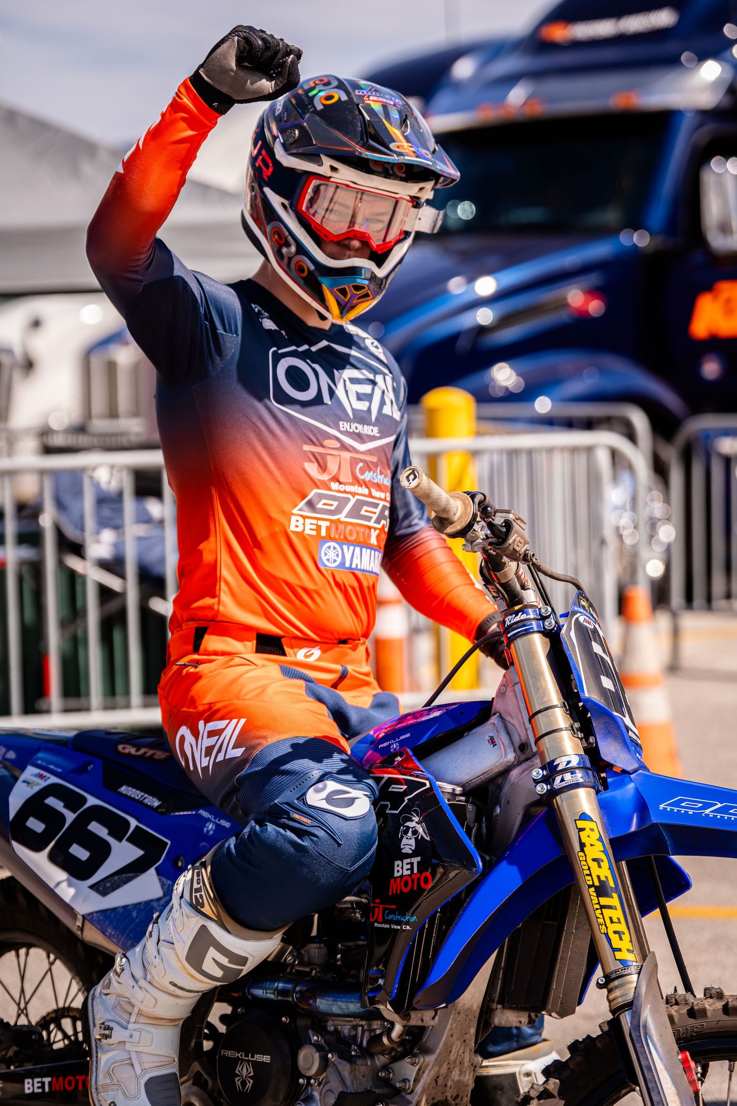Dirt bike rider in orange gear mounted on blue motorcycle in racing paddock.