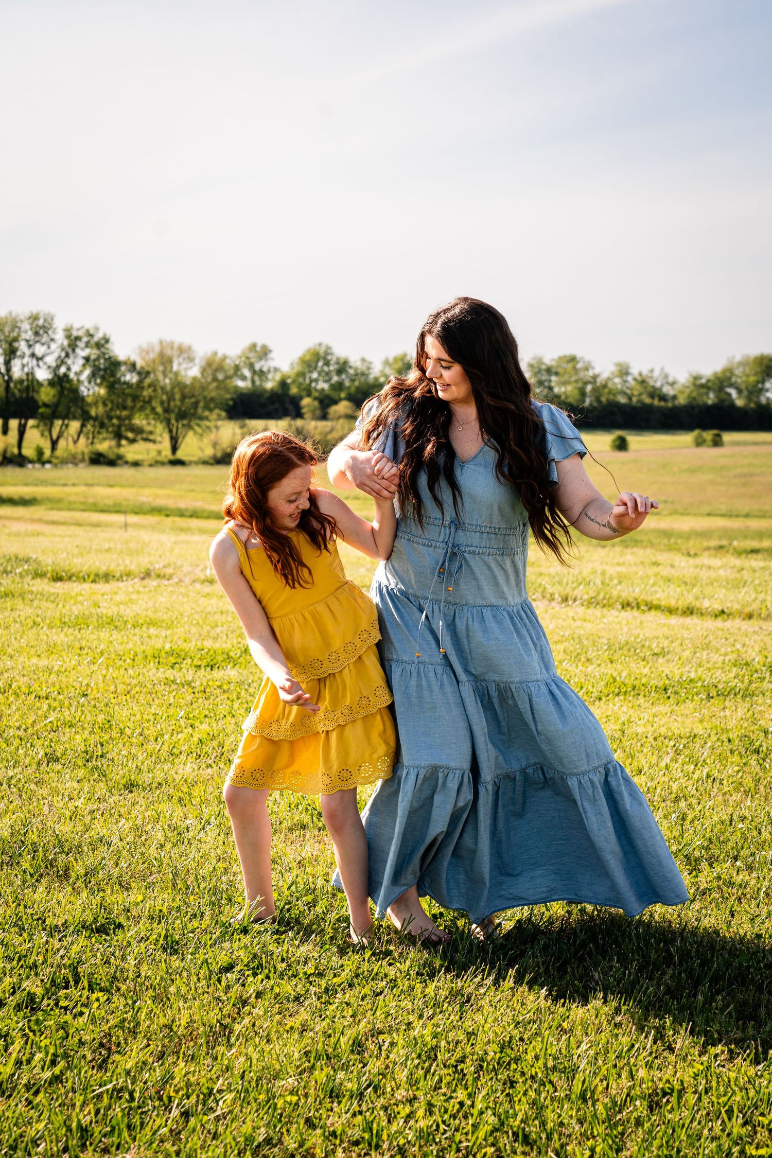 Two people in flowing dresses dance joyfully together in a sunlit summer field.