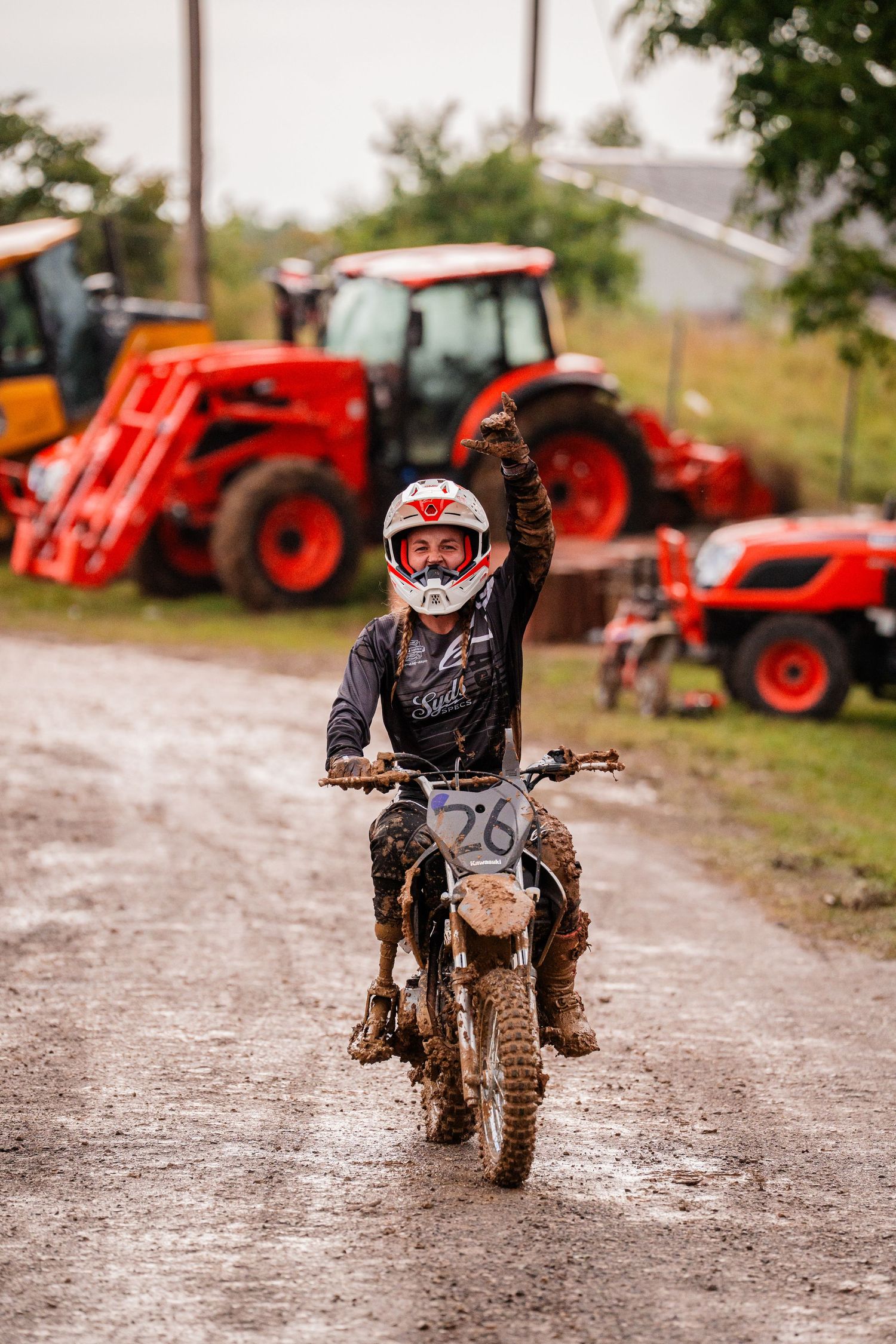 Dirt bike rider speeding down gravel path with red tractors in background.