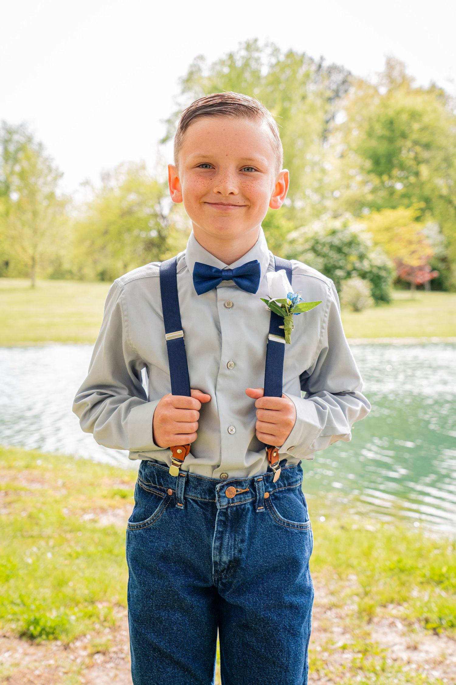 Young attendant in formal attire with suspenders and bow tie poses by a pond on sunny day.