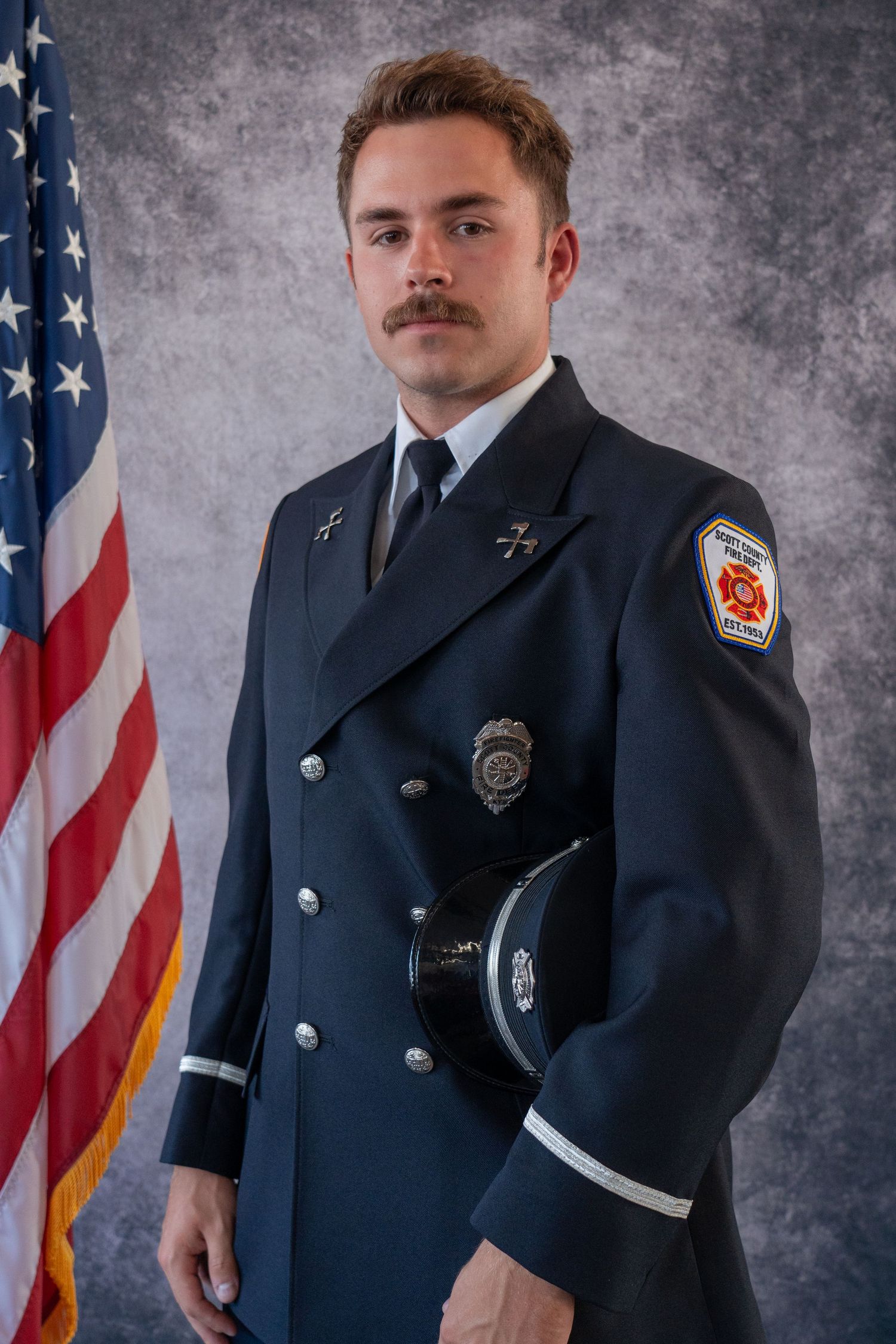 Uniformed firefighters in formal dress standing at attention with American flag backdrop.