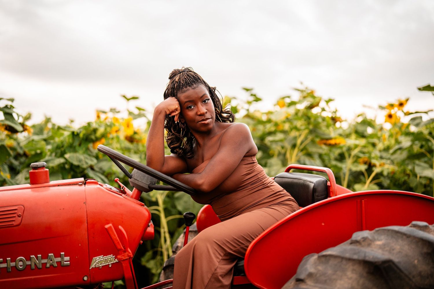 A woman in a brown dress sitting on a red tractor in a field of sunflowers.