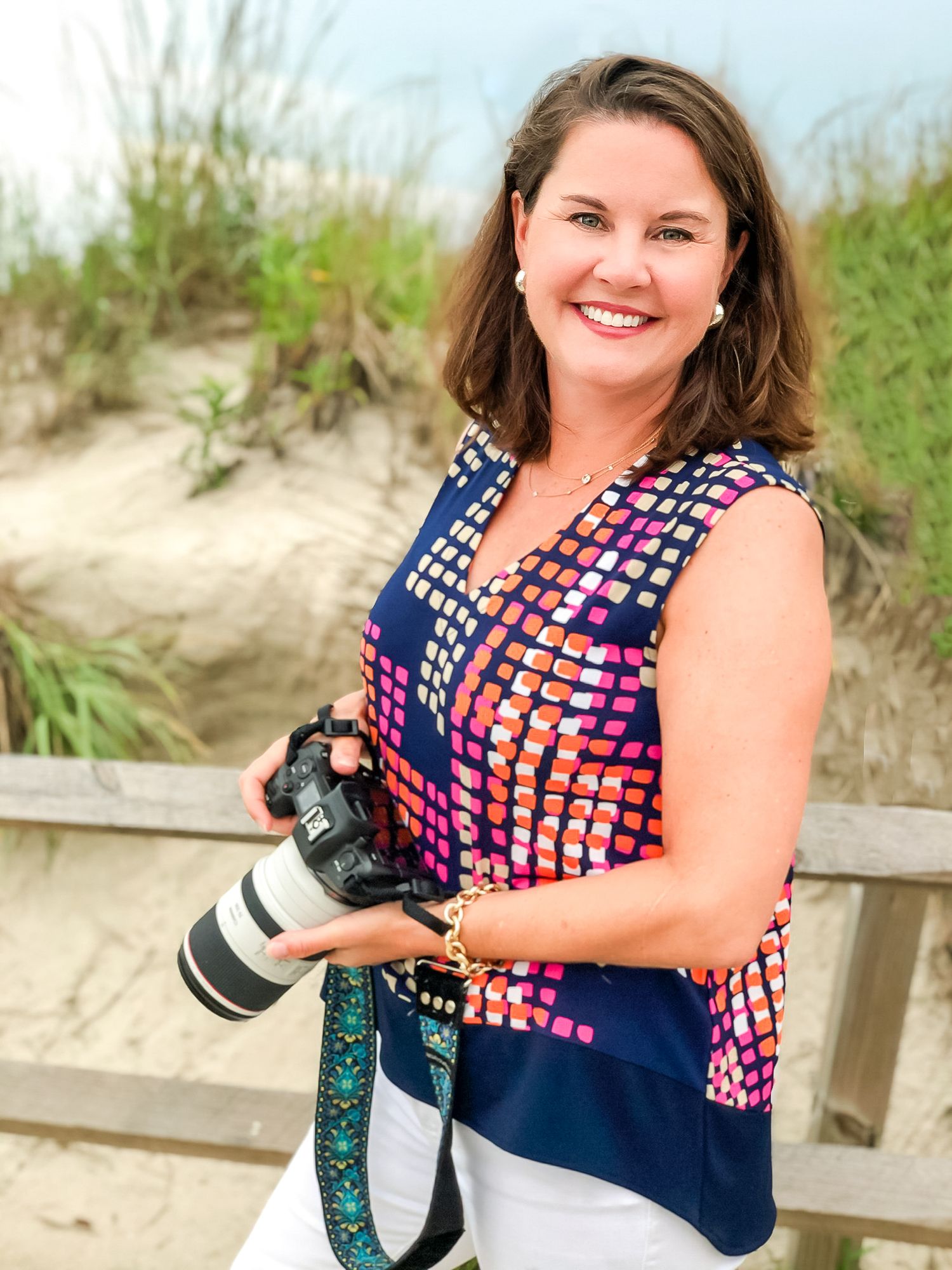 A photographer in a navy and pink patterned top smiles while holding a professional camera on a beach boardwalk.