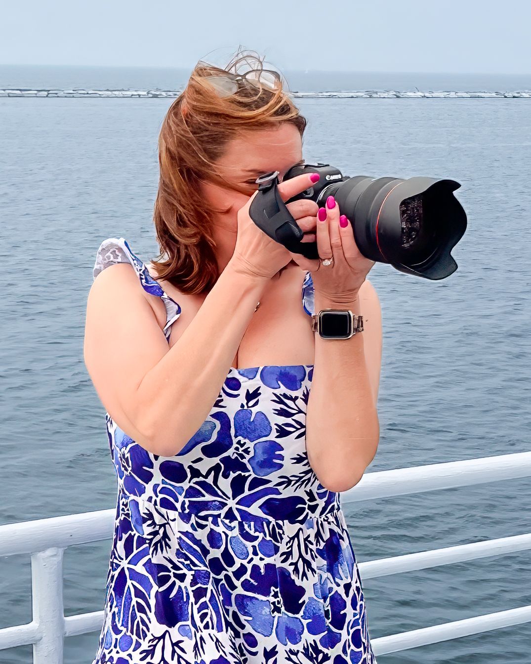 A photographer in a blue and white floral dress takes photos from a boat deck with a professional camera and telephoto lens.