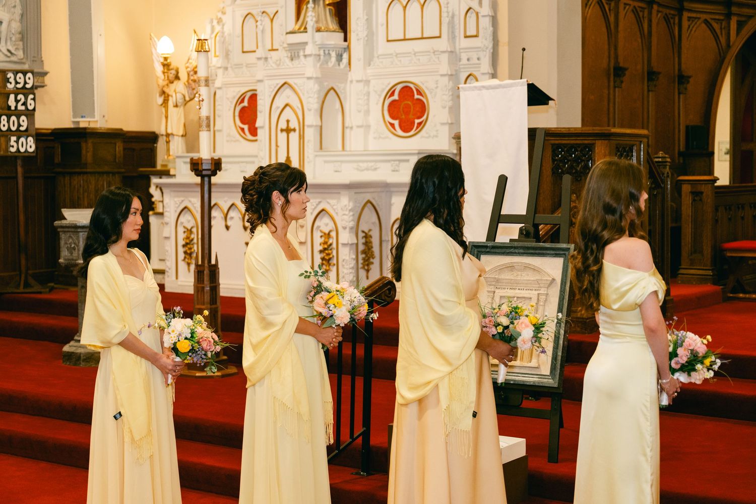 Bridesmaids in yellow gowns holding bouquets stand at church altar during wedding ceremony.