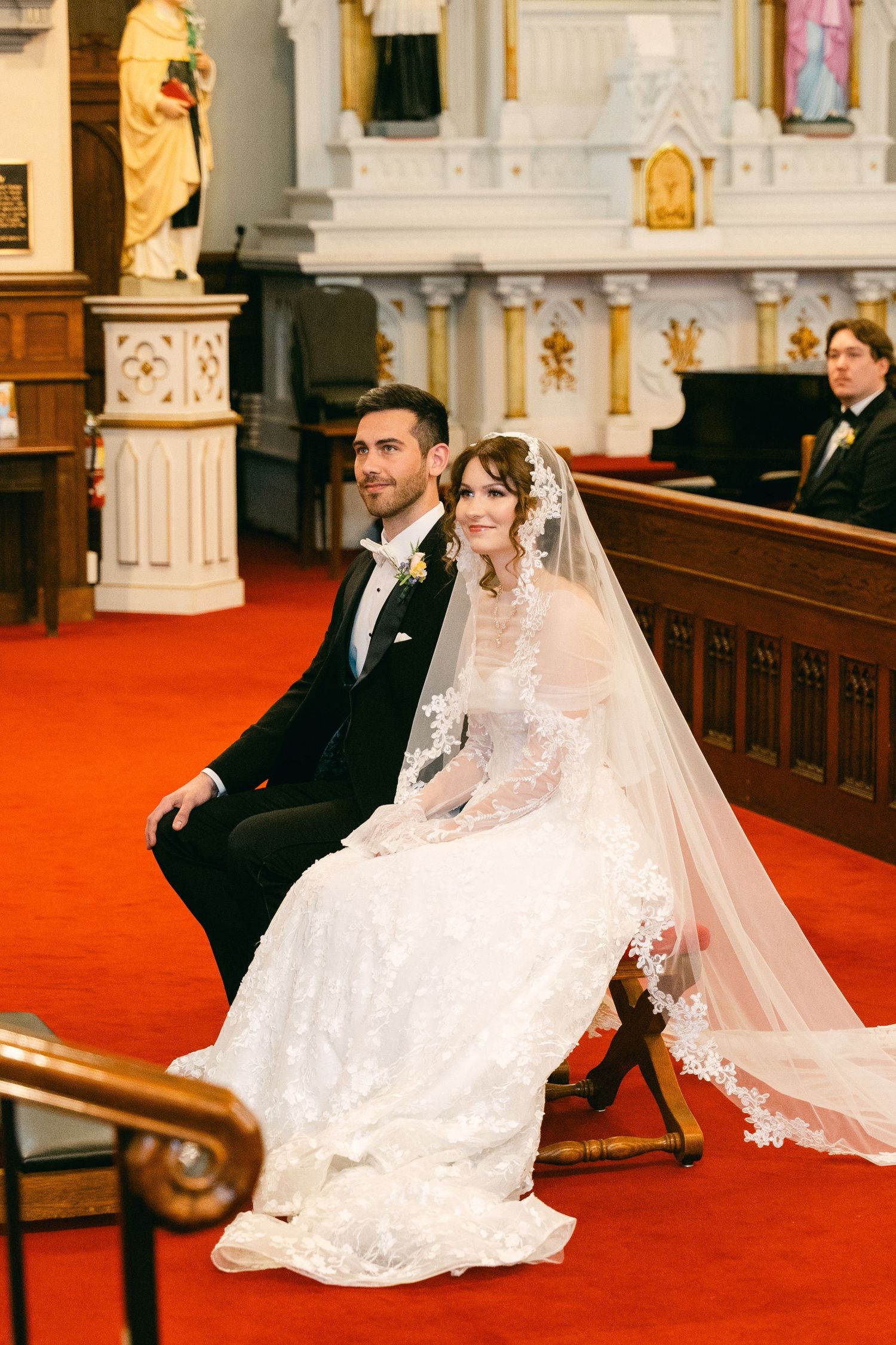 Bride and groom seated together during church wedding ceremony, bride wearing lace gown and veil on red carpet aisle.