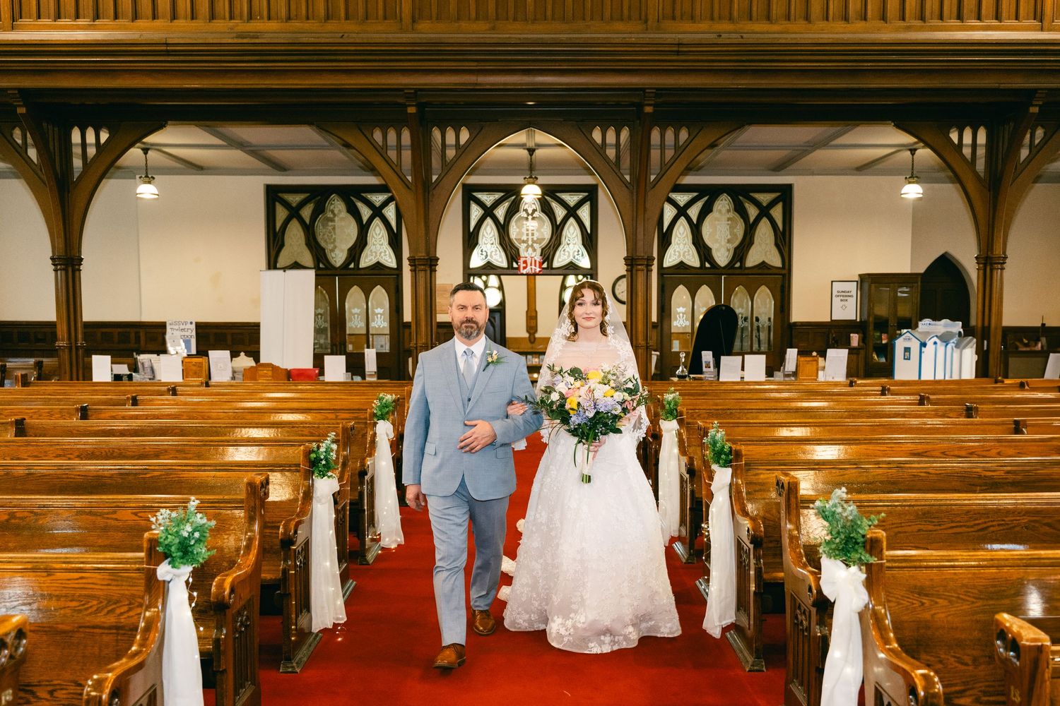 Bride and groom walking down red carpet aisle in elegant wooden church decorated with white bows and greenery.
