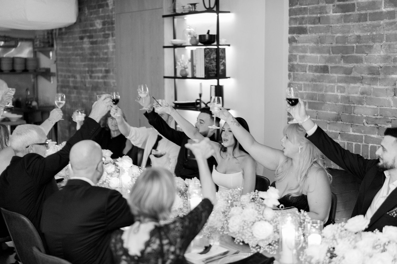 Wedding guests raise their glasses in a celebratory toast at a reception with exposed brick walls.