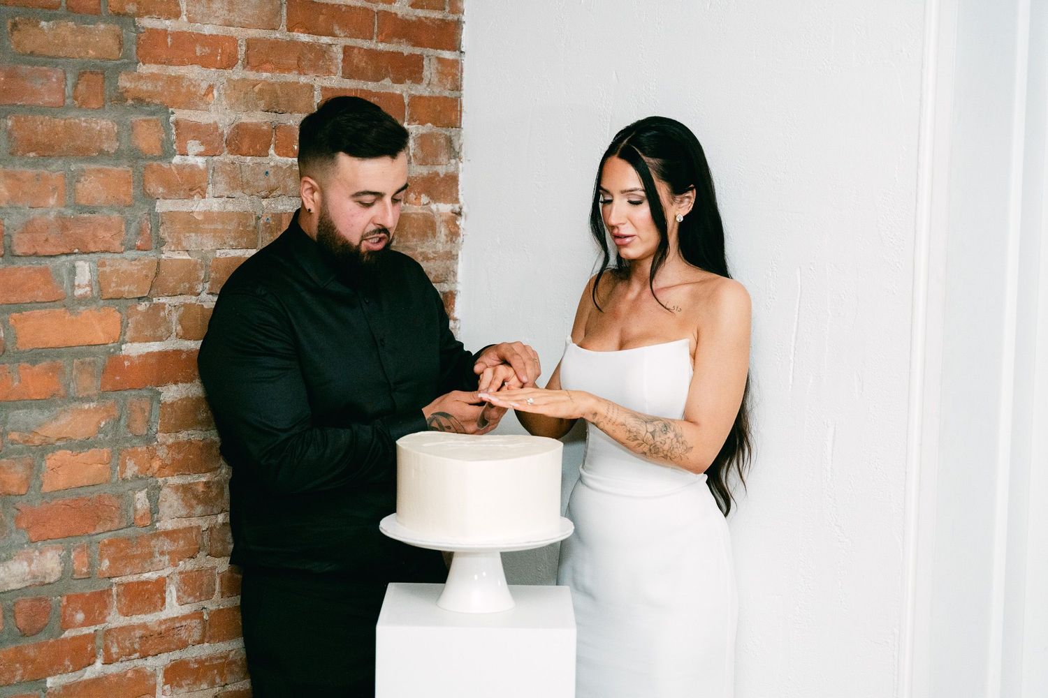 Couple in wedding attire sharing cake together against exposed brick wall backdrop.