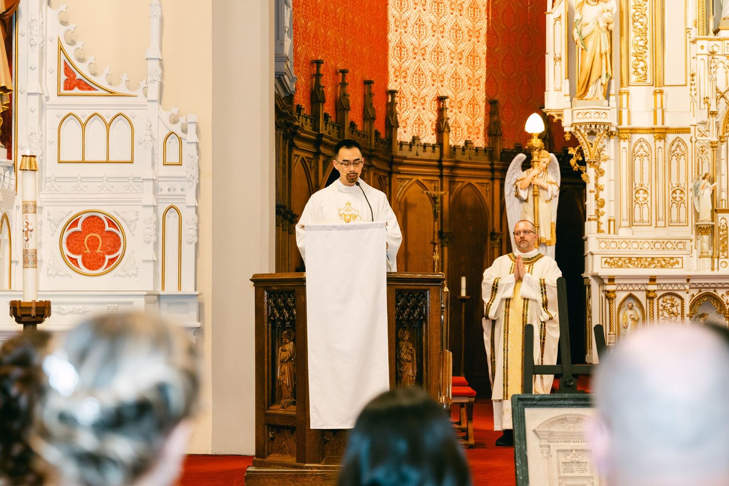 Catholic priests in white vestments celebrate Mass at an ornate church altar during a religious ceremony.