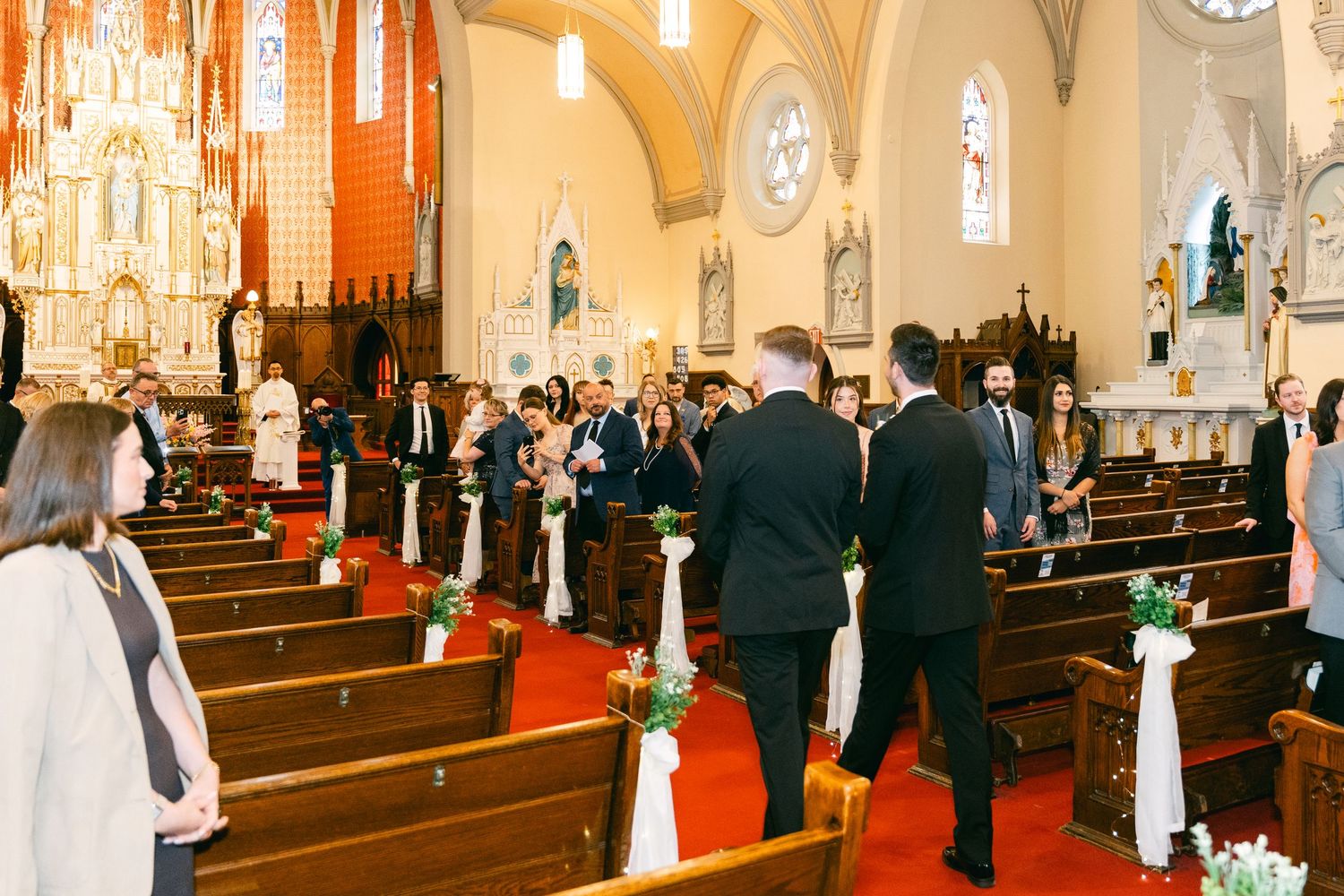 Wedding party processional down church aisle with floral pew decorations and stained glass windows.