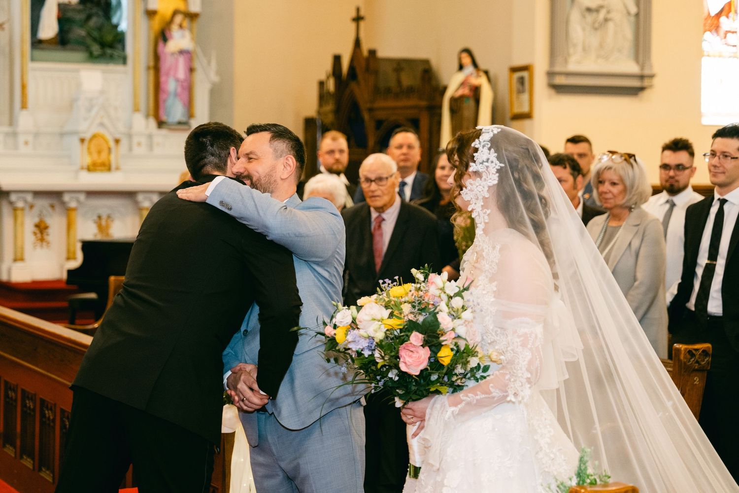 Bride in white lace gown holding floral bouquet greeted by guests at church wedding ceremony altar.