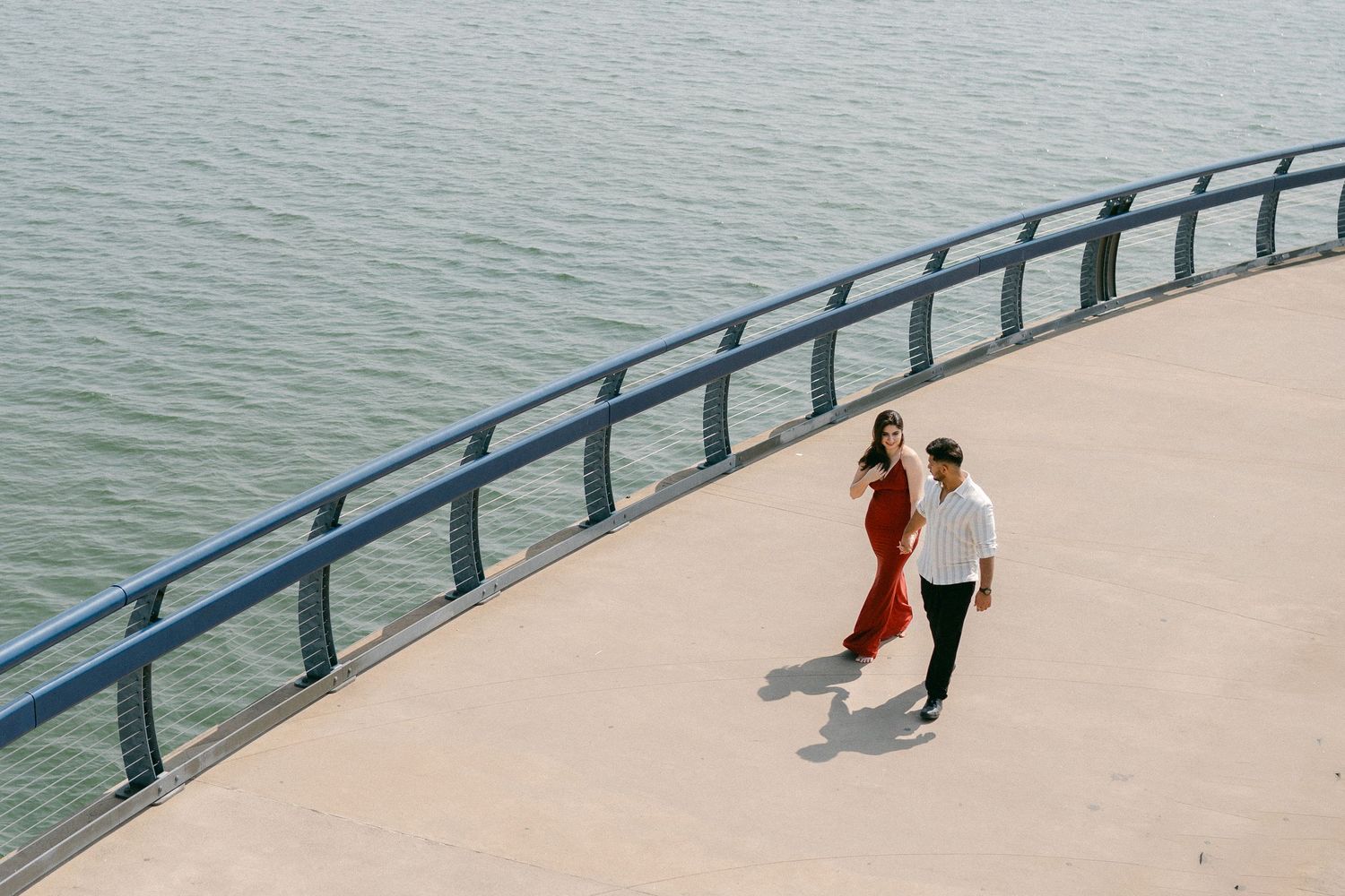 A couple in casual attire walking along a curved waterfront path with metal railings.